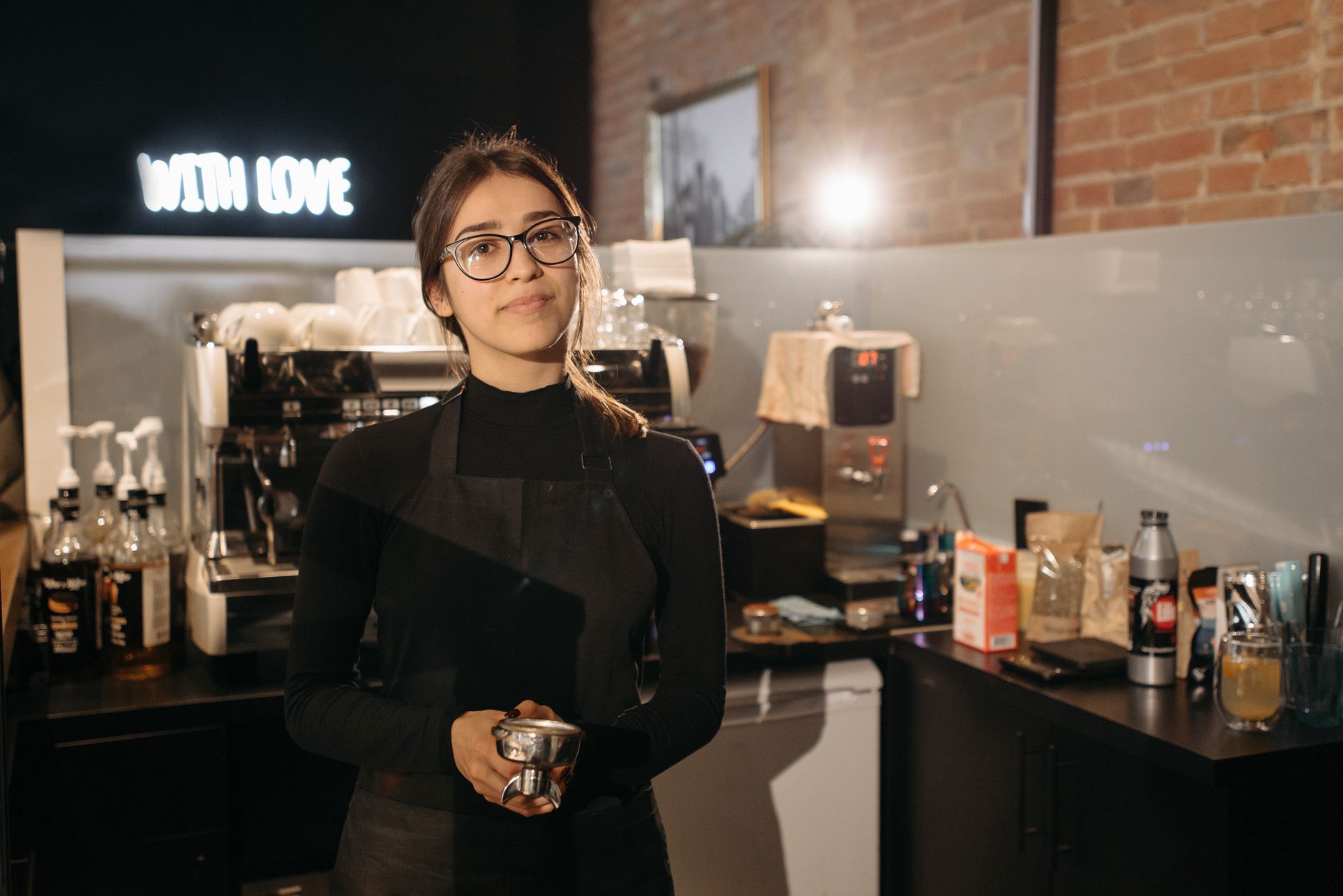 Smiling barista holding portafilter inside a cozy cafe with coffee machine, wearing apron and glasses.