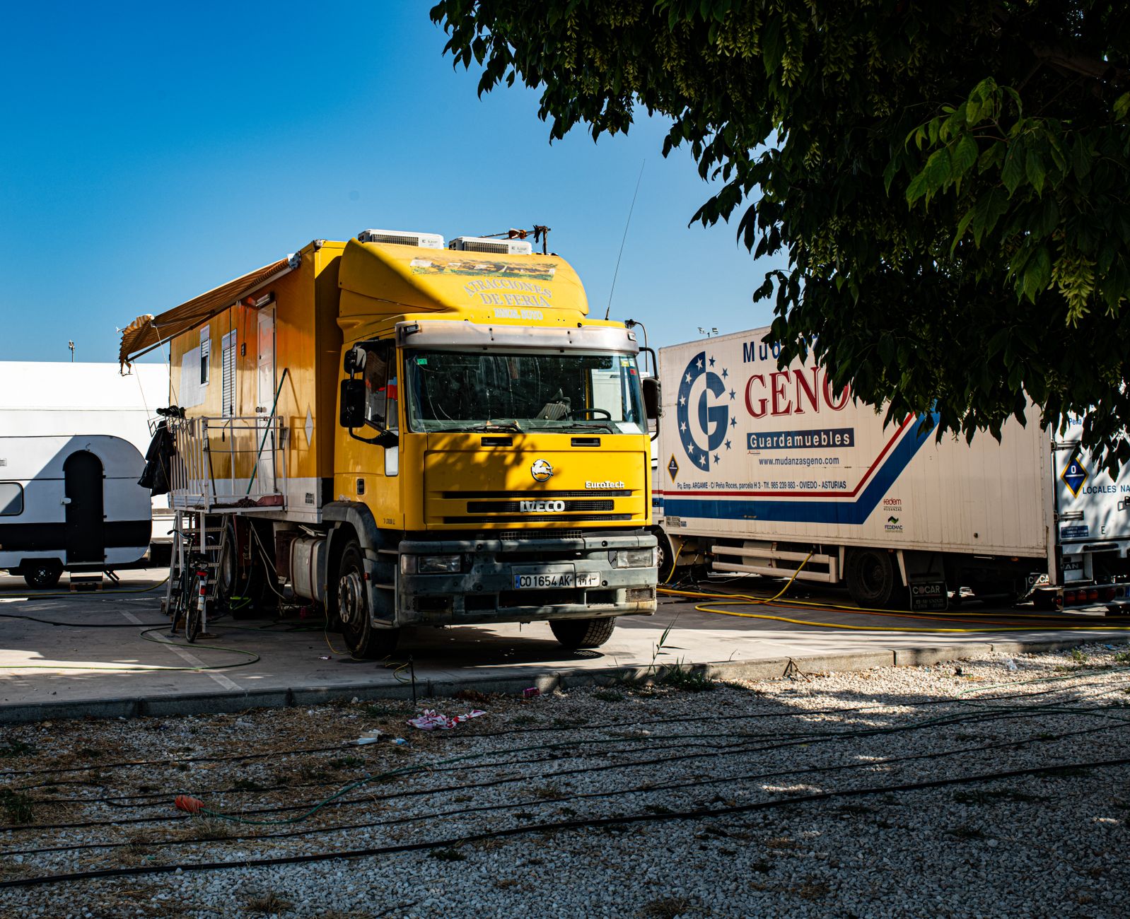 A vibrant yellow truck is parked in an outdoor industrial area under a tree.