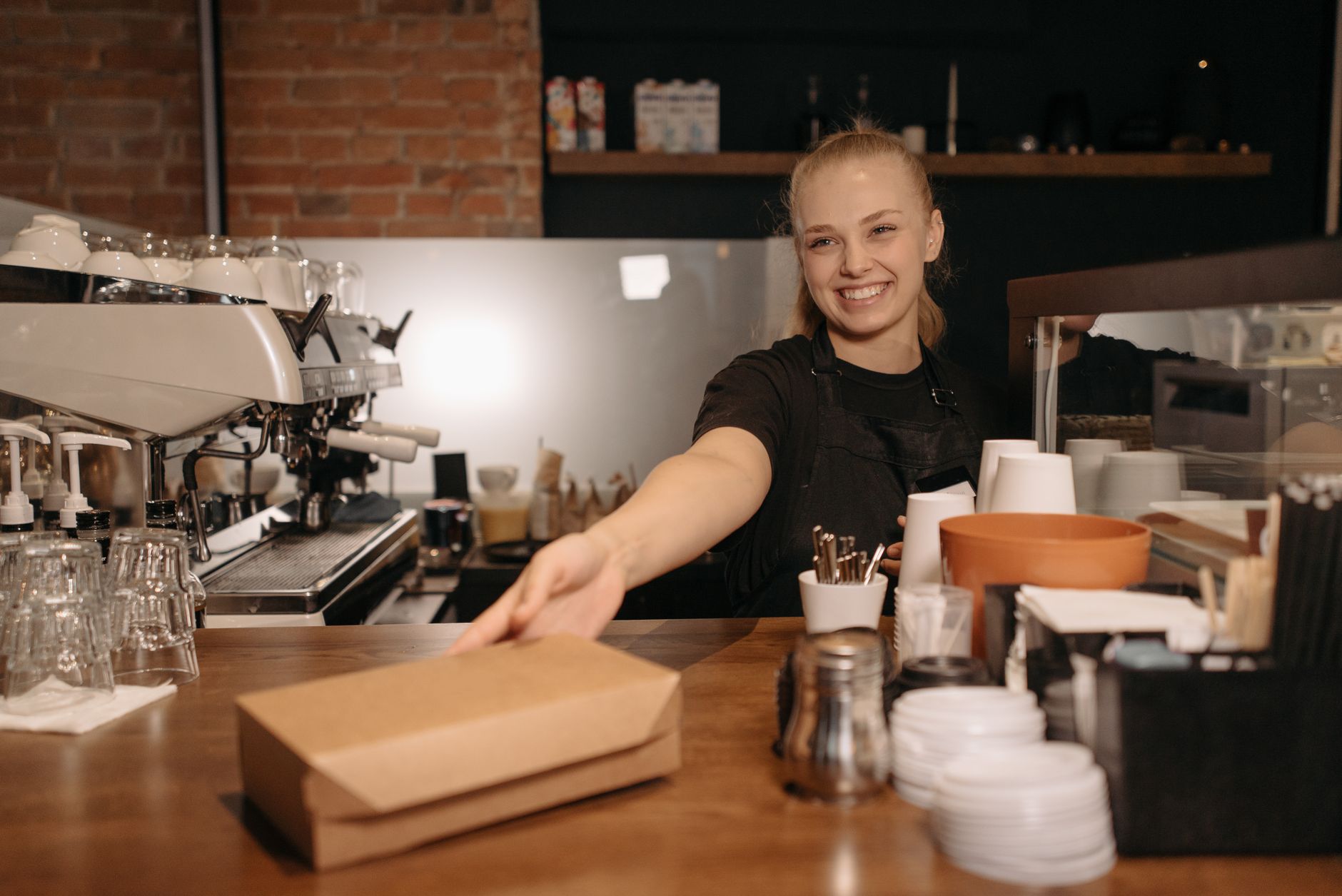 Smiling barista handing a takeout box at a modern coffee shop counter.