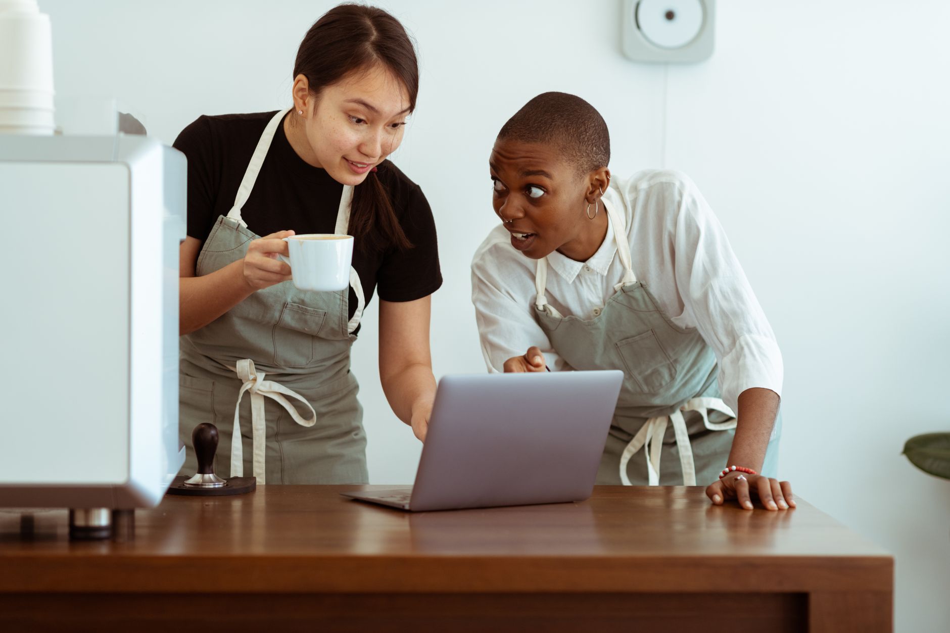 Cheerful surprised diverse colleagues in aprons using modern netbook and sharing business ideas while gathering at table with cup of drink in light cozy kitchen