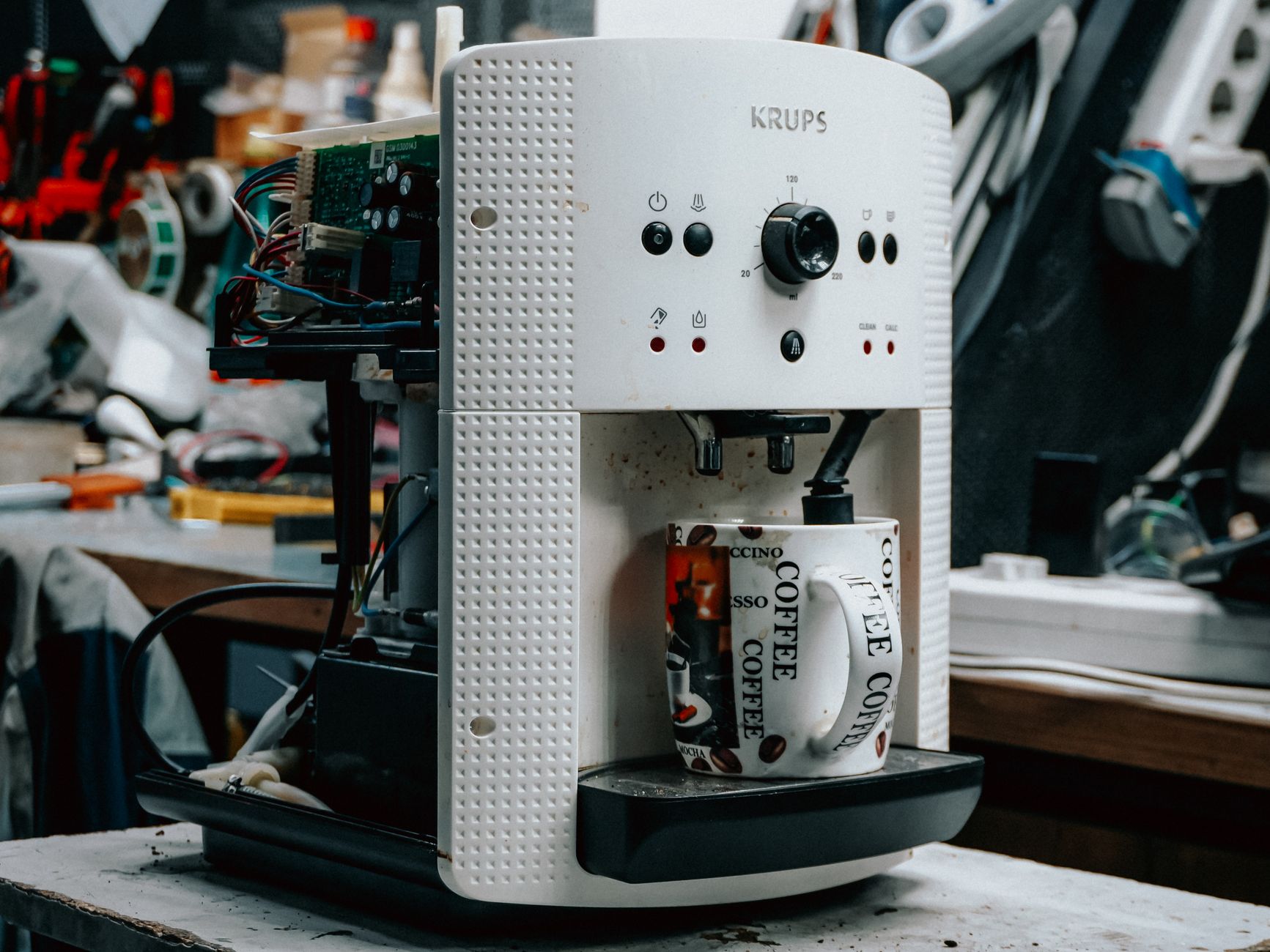 A coffee machine undergoing repair in a cluttered workshop, with parts exposed and a coffee mug.