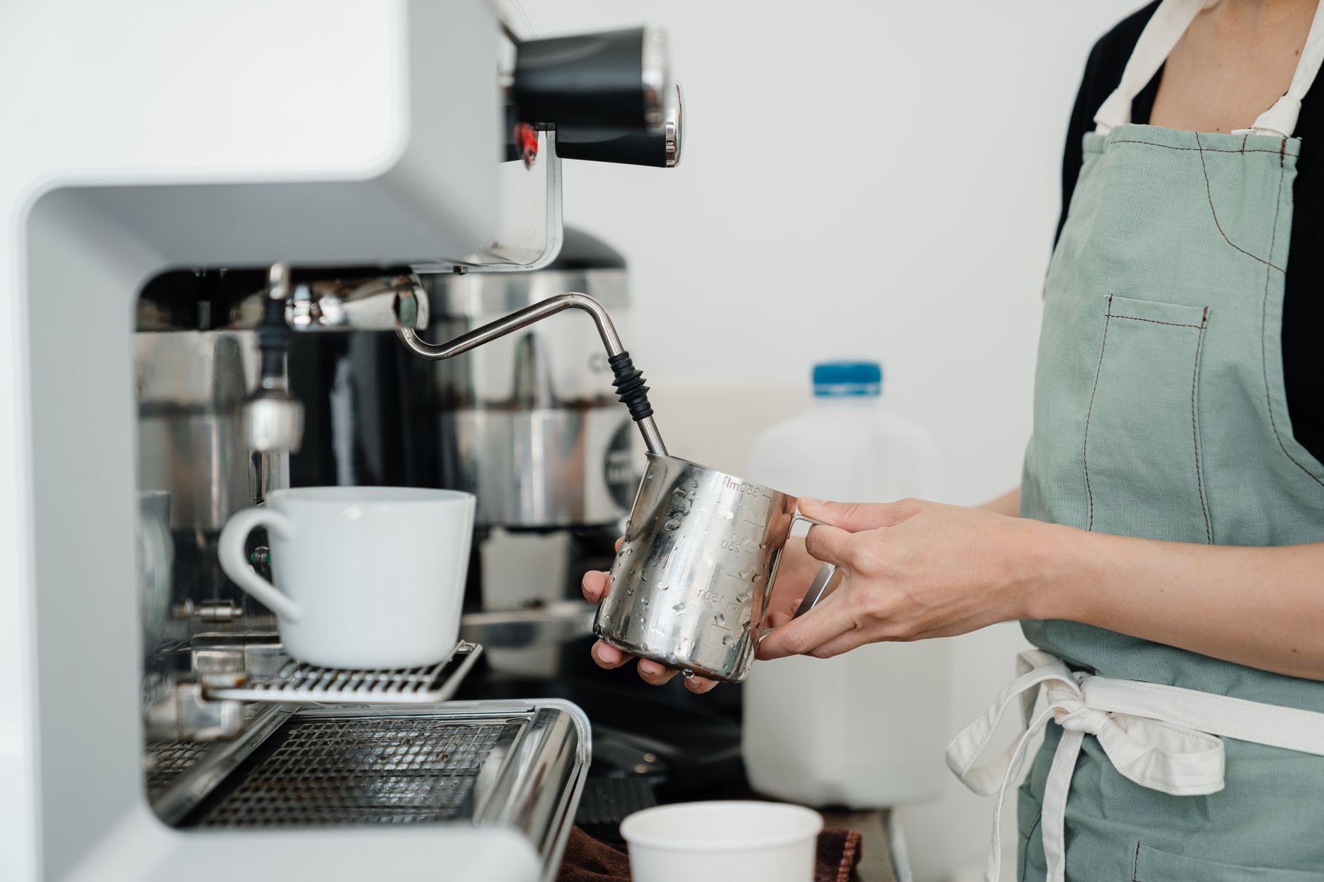 Crop unrecognizable young female barista in apron heating milk in pitcher with modern coffee machine during work in cozy cafe