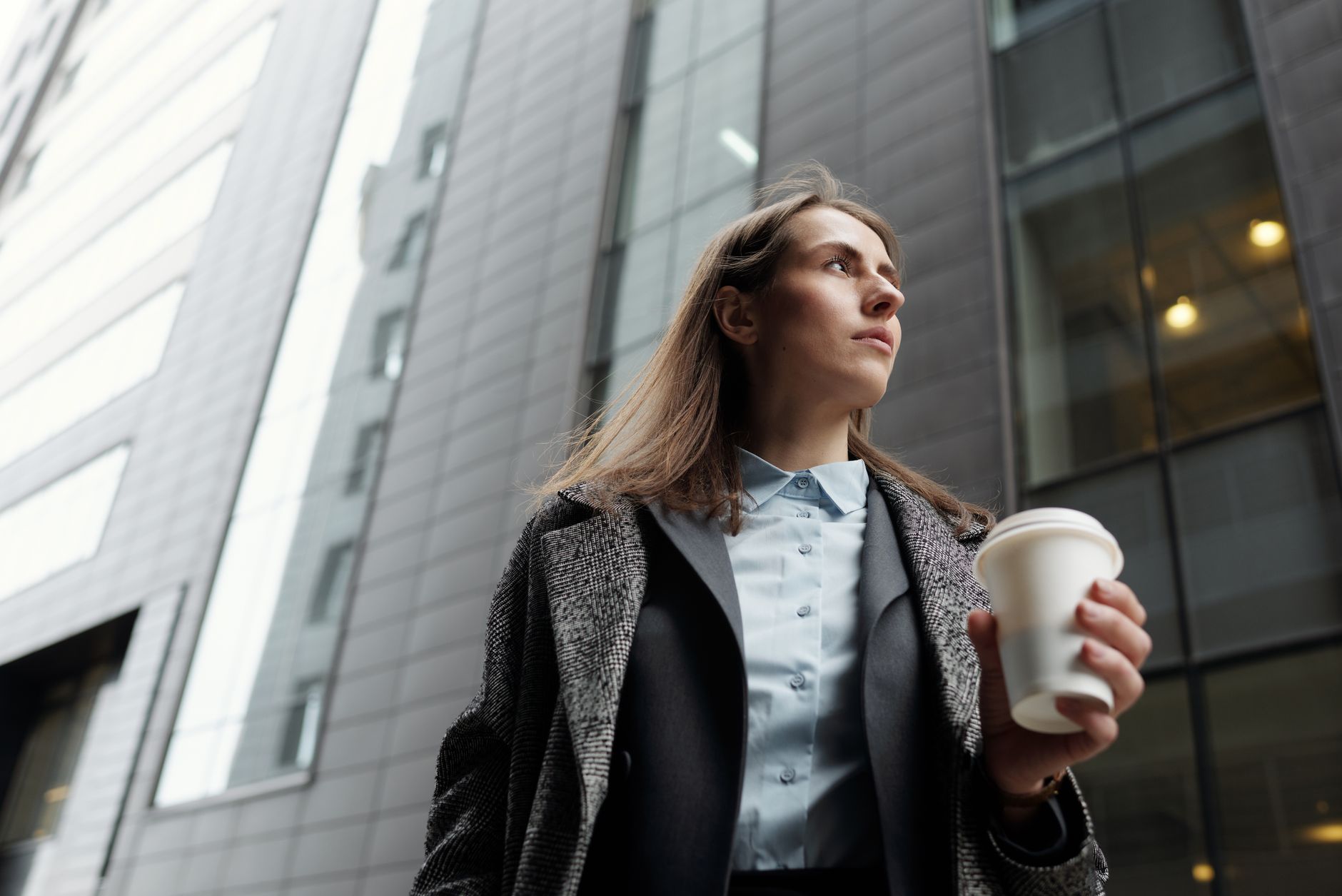 Professional woman in a city street holding a coffee cup, gazing upwards.
