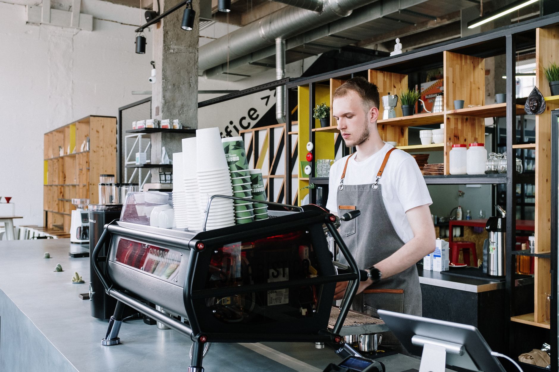 A young male barista prepares coffee in a trendy, open-space café, surrounded by modern equipment.