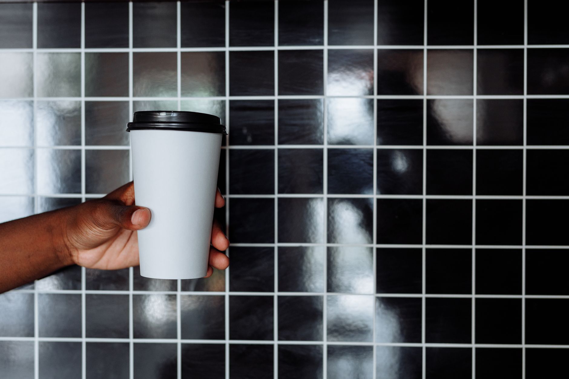 A hand holding a white disposable coffee cup against a tiled black background.