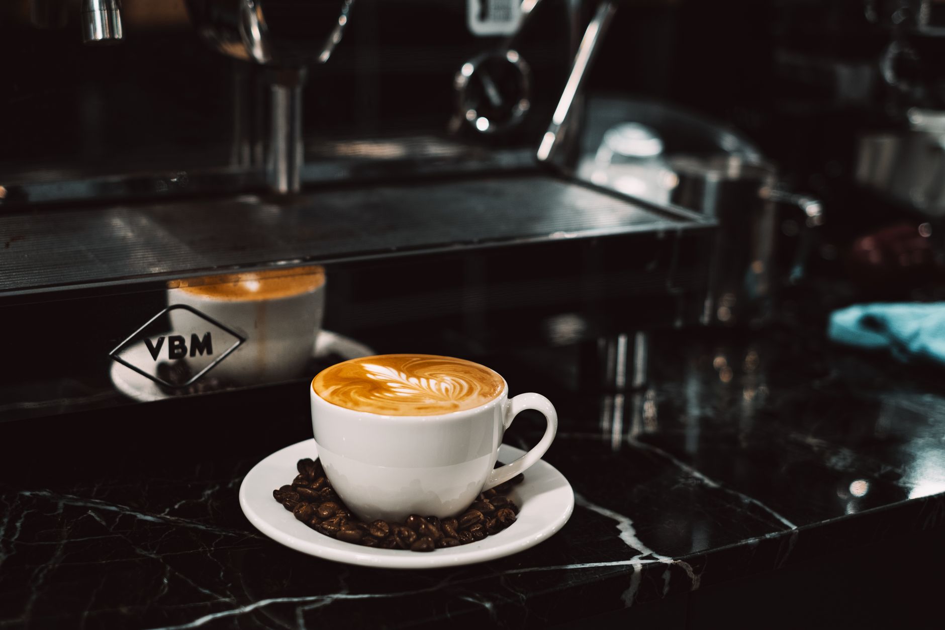 A beautifully crafted cappuccino with intricate latte art sits on a marble counter in a cozy coffee shop.