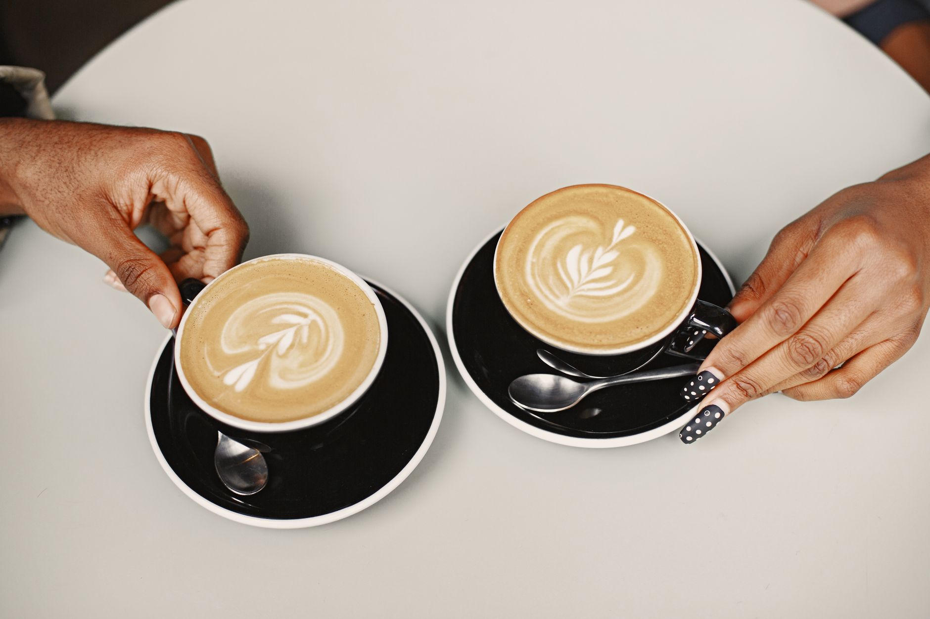 Close-up of two people holding lattes with intricate foam art designs in black cups and saucers.