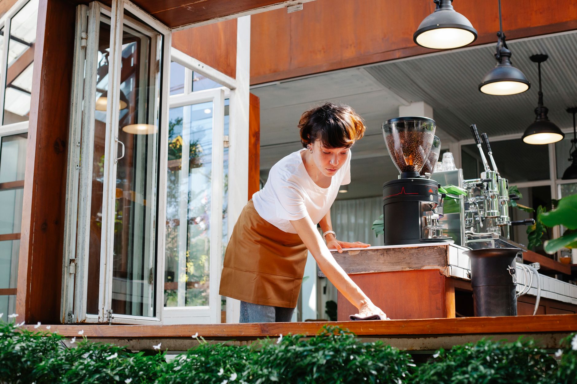 From below of focused female in apron cleaning wooden counter of modern coffee house with coffee makers and special equipment