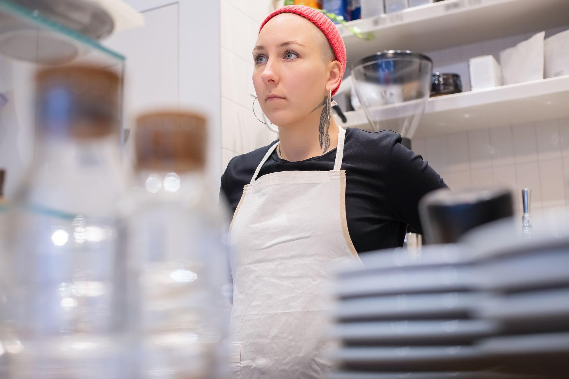 Female barista wearing apron and beanie in a stylish coffee shop setting.