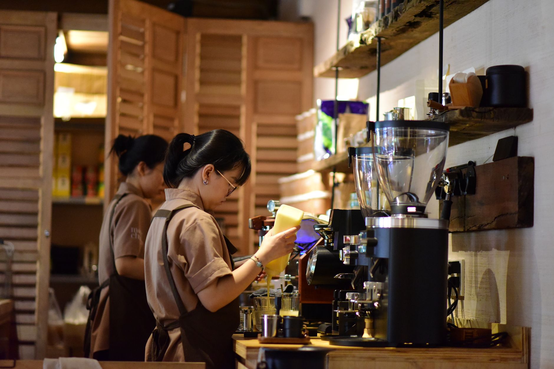 Two baristas prepare coffee behind the counter in a warm, inviting cafe setting.