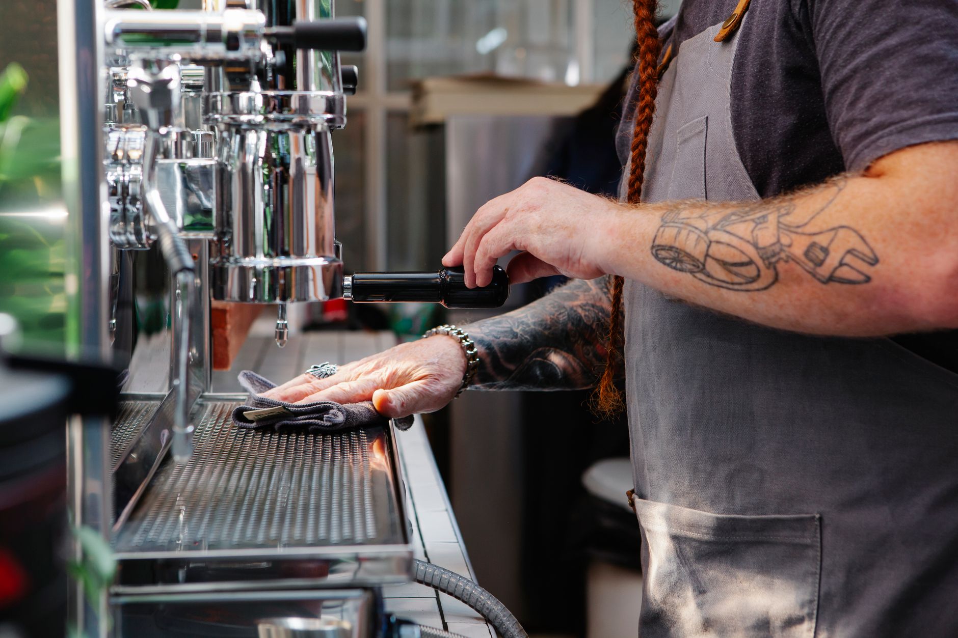 Side view of crop anonymous tattooed male employee cleaning professional coffee maker with rag in cafeteria kitchen