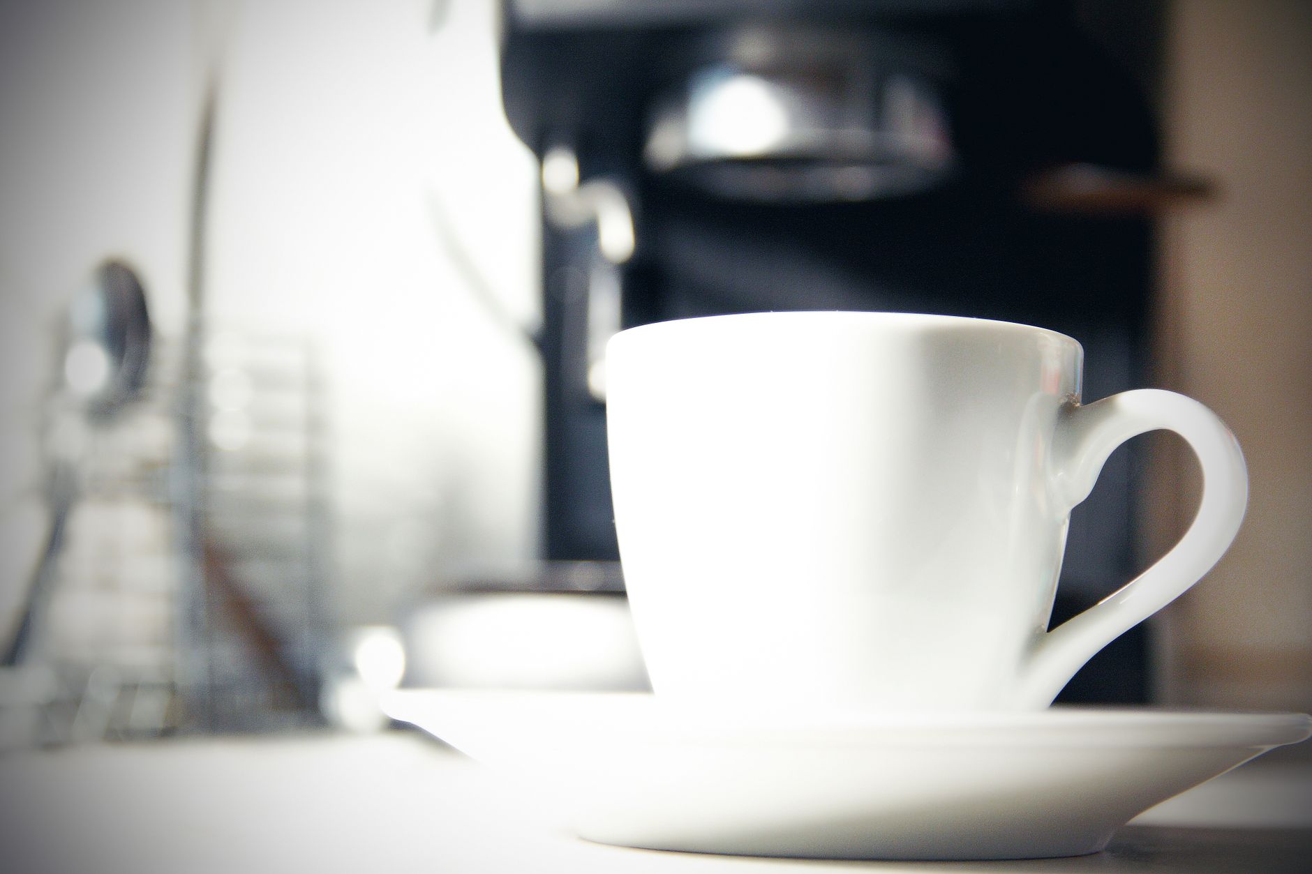 A white coffee cup on saucer with a blurred background of a coffee machine in a kitchen setting.