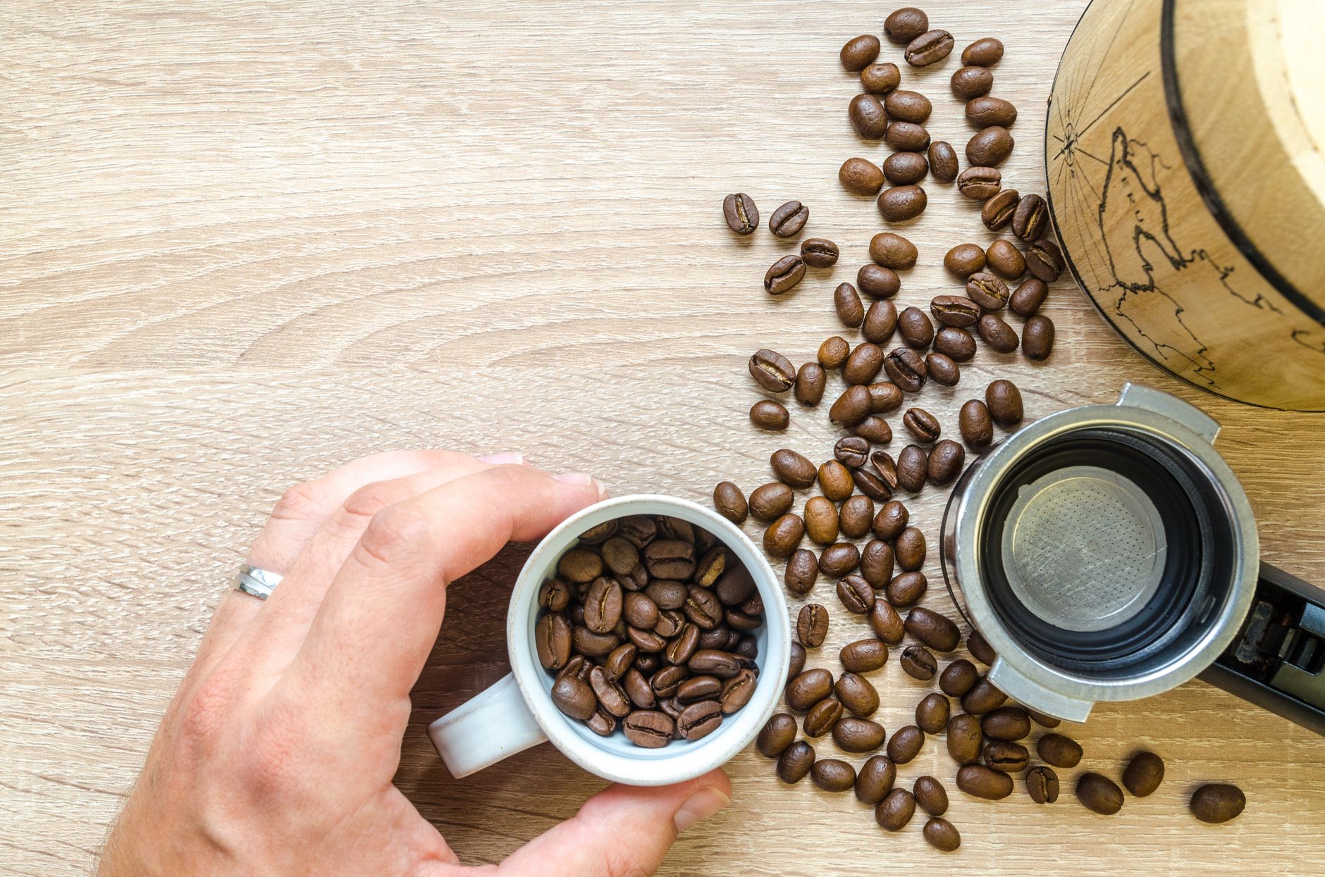 A hand holds a cup filled with roasted coffee beans beside a coffee maker on a wooden table.