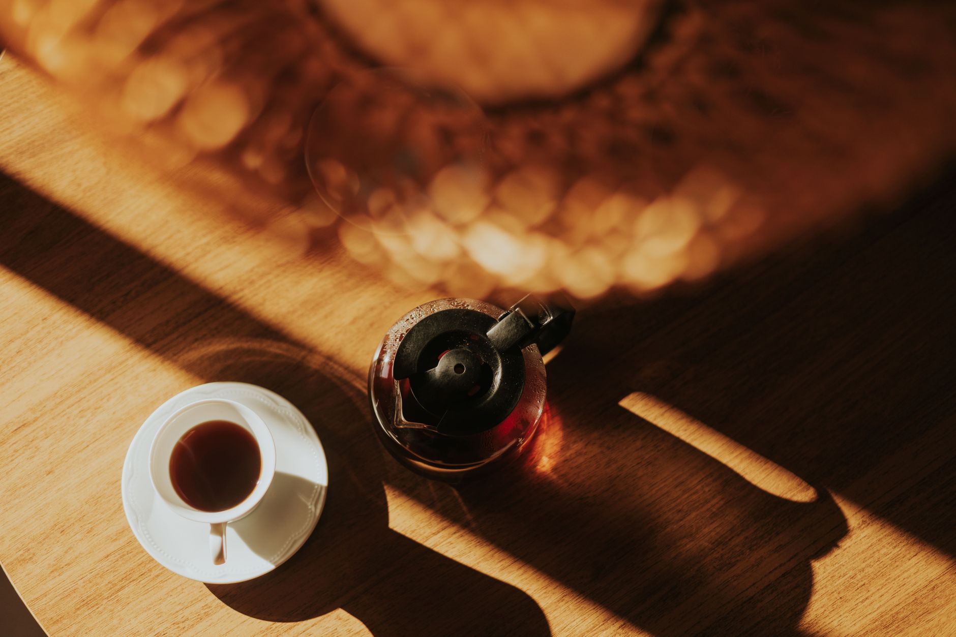 Warm sunlight casts shadows on a table with a coffee cup and French press.