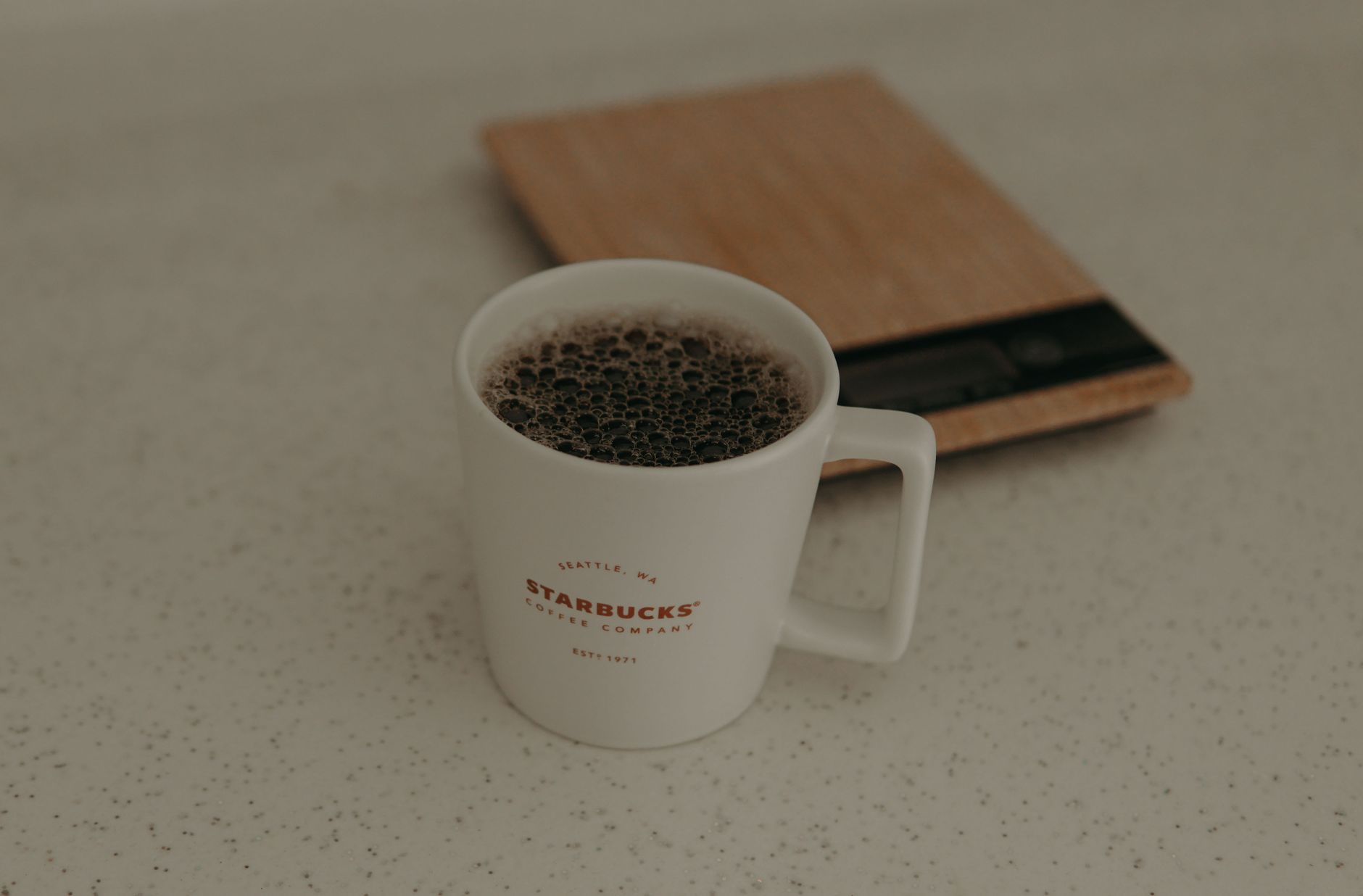 A close-up of a Starbucks coffee mug filled with coffee next to a weighing scale on a speckled surface.