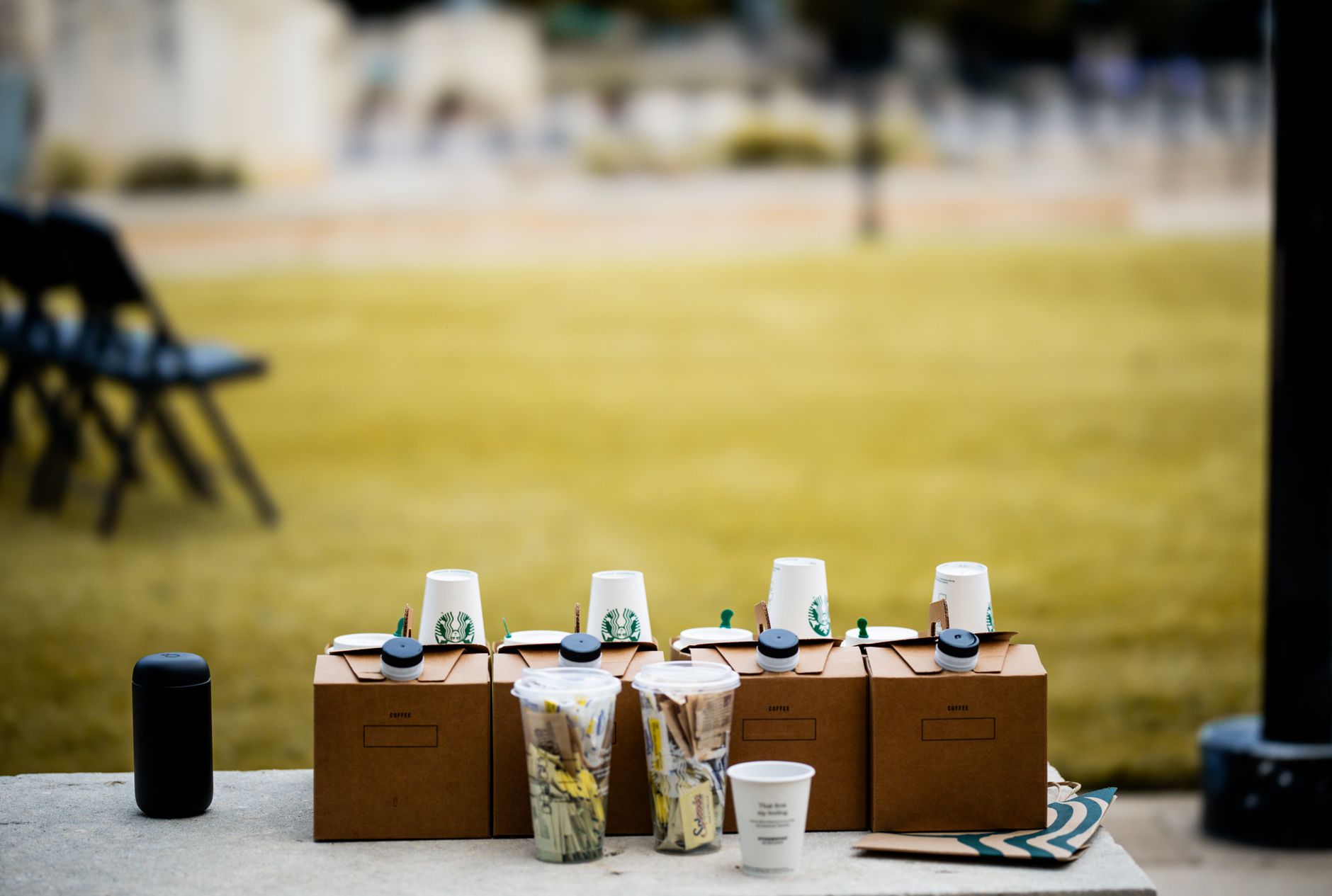 A row of coffee containers with cups and supplies set outdoors at an event.