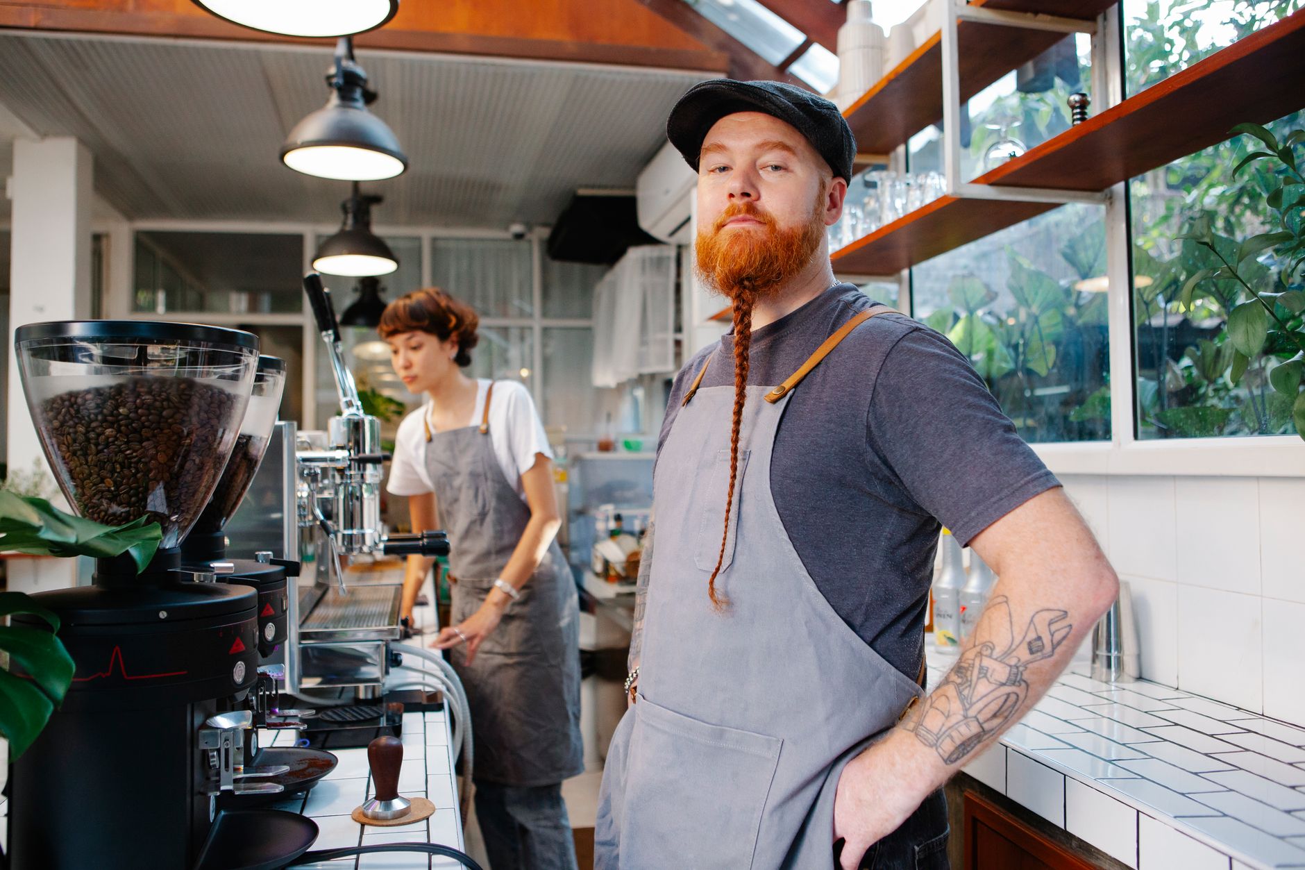 Young bearded male looking at camera while preparing coffee with colleague in kitchen with coffee machines in cafe