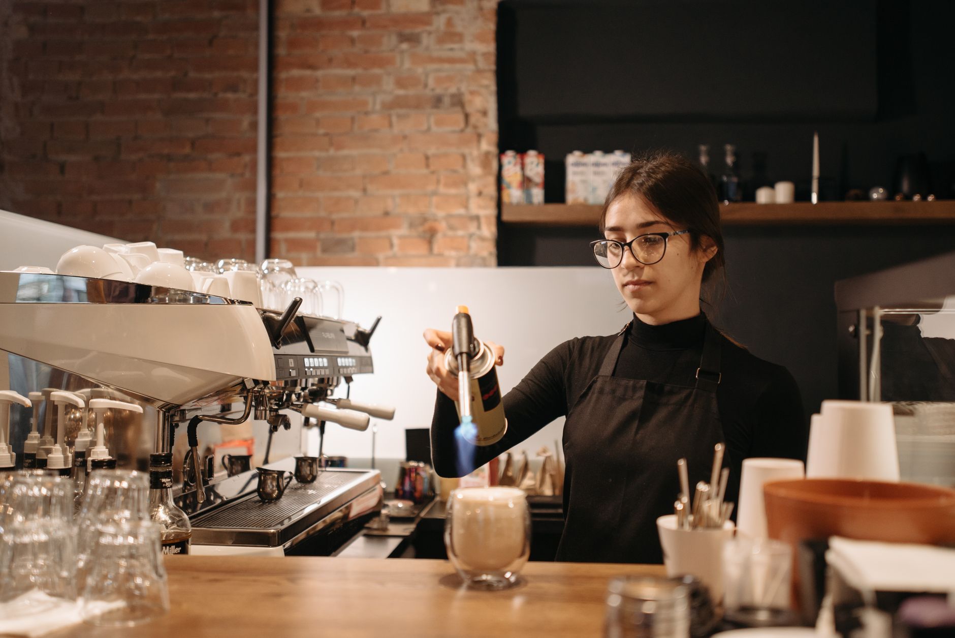 Barista using a torch to prepare a coffee drink in a cozy, brick-exposed cafe.