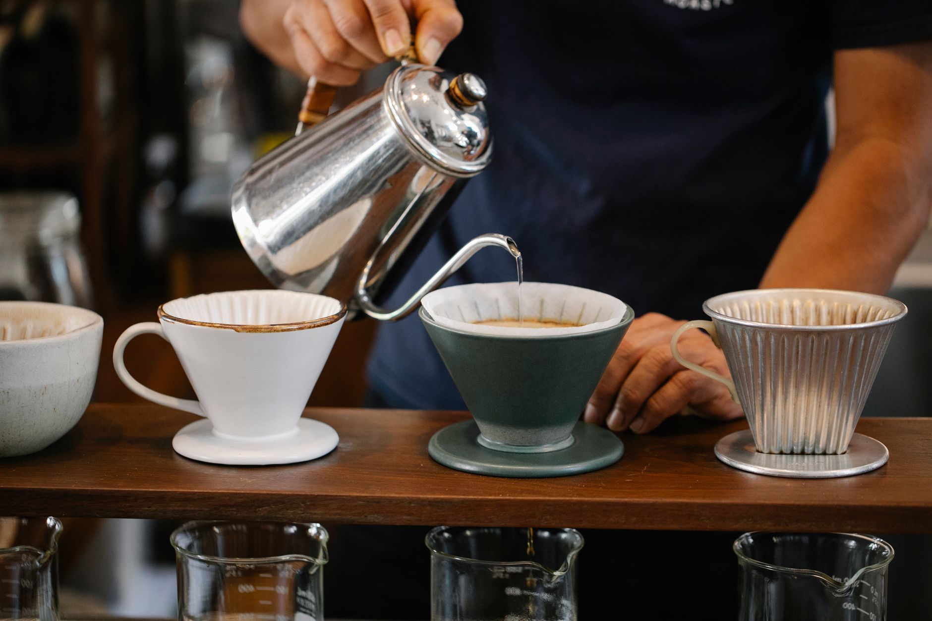 Crop anonymous barista pouring hot water from coffee kettle into drippers on wooden stand while making pour over coffee in cafe on blurred background