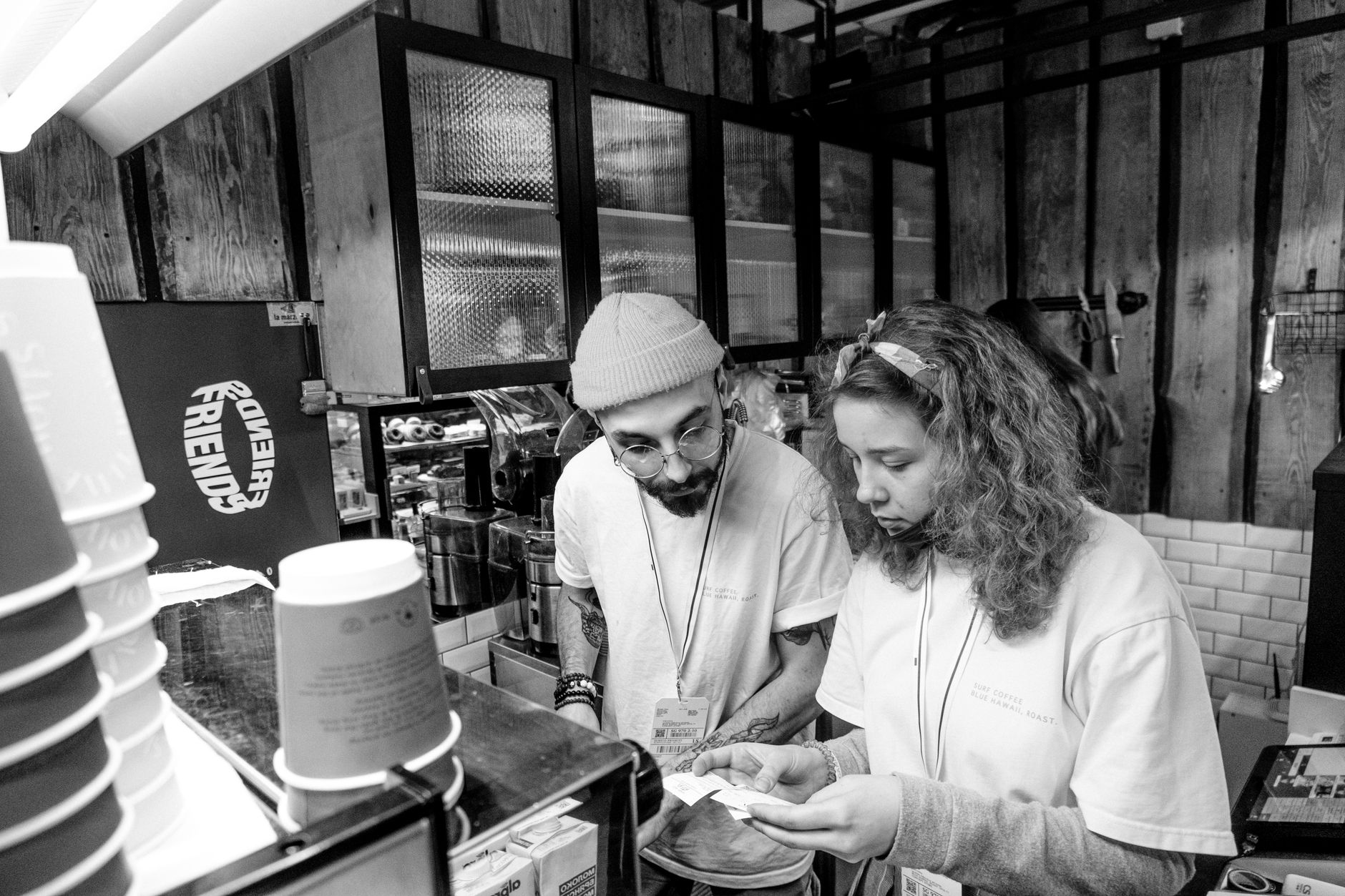 Two baristas in a coffee shop reviewing an order, wearing t-shirts and eyeglasses.