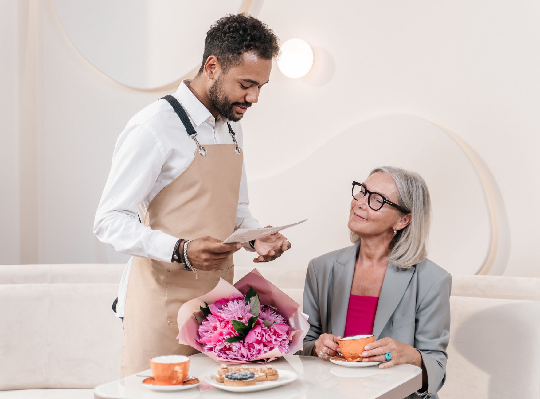 A waiter delivers a floral bouquet to a woman at a table in a stylish café setting.