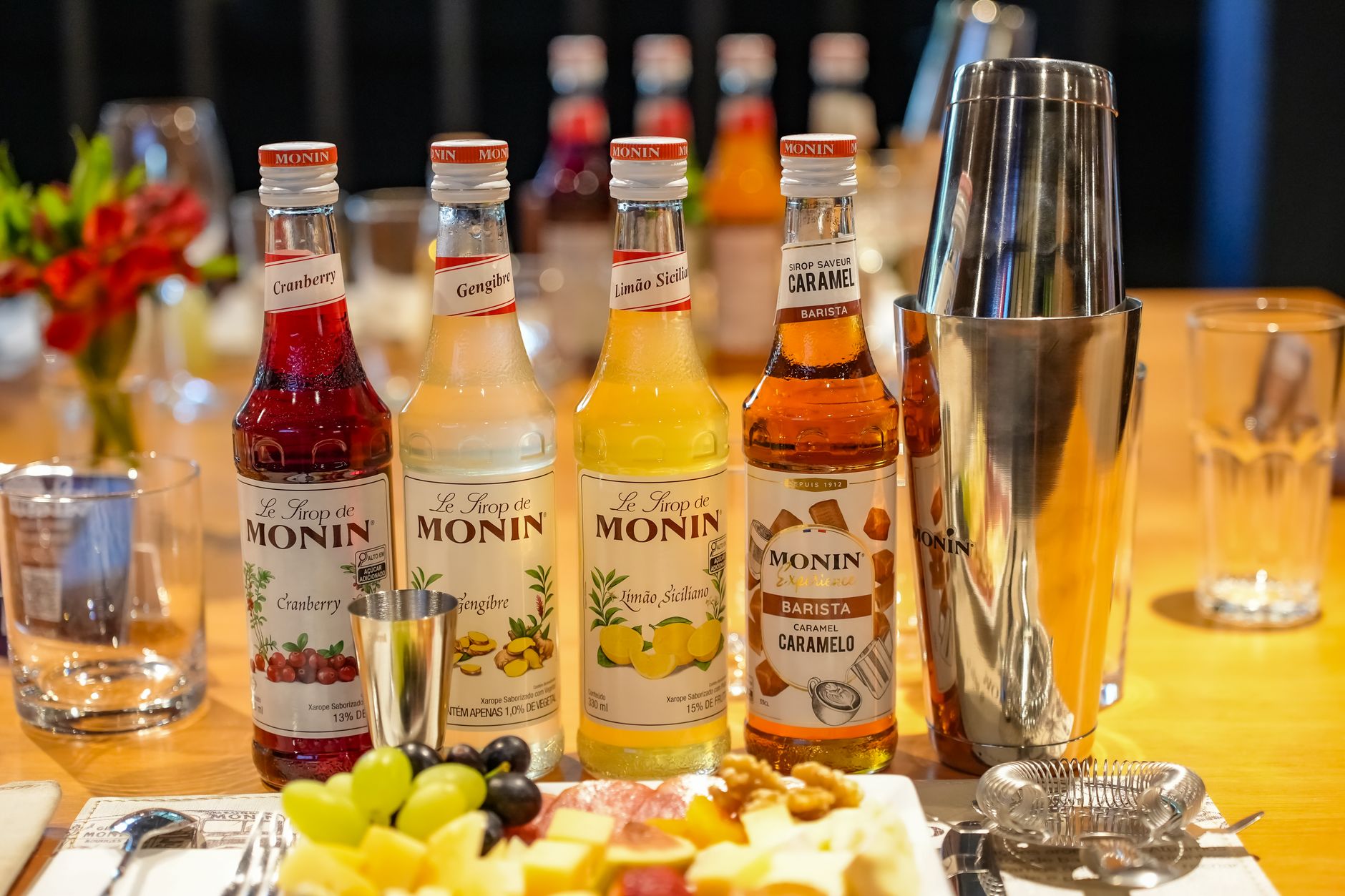 Vibrant syrup bottles from Monin next to a cocktail shaker, surrounded by fresh fruit and glasses, ideal for bar settings.