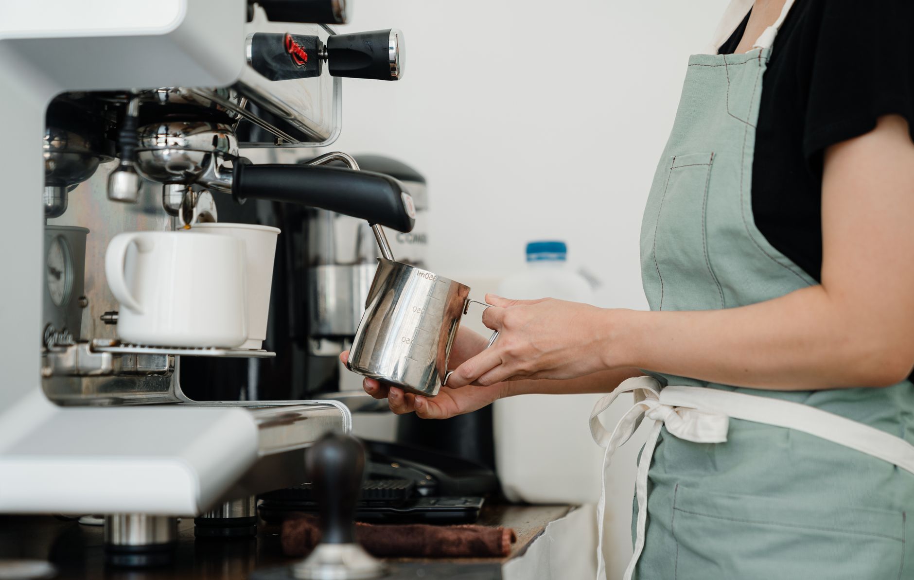 Crop side view of unrecognizable young woman in apron steaming milk in steel pot using professional coffee maker at counter