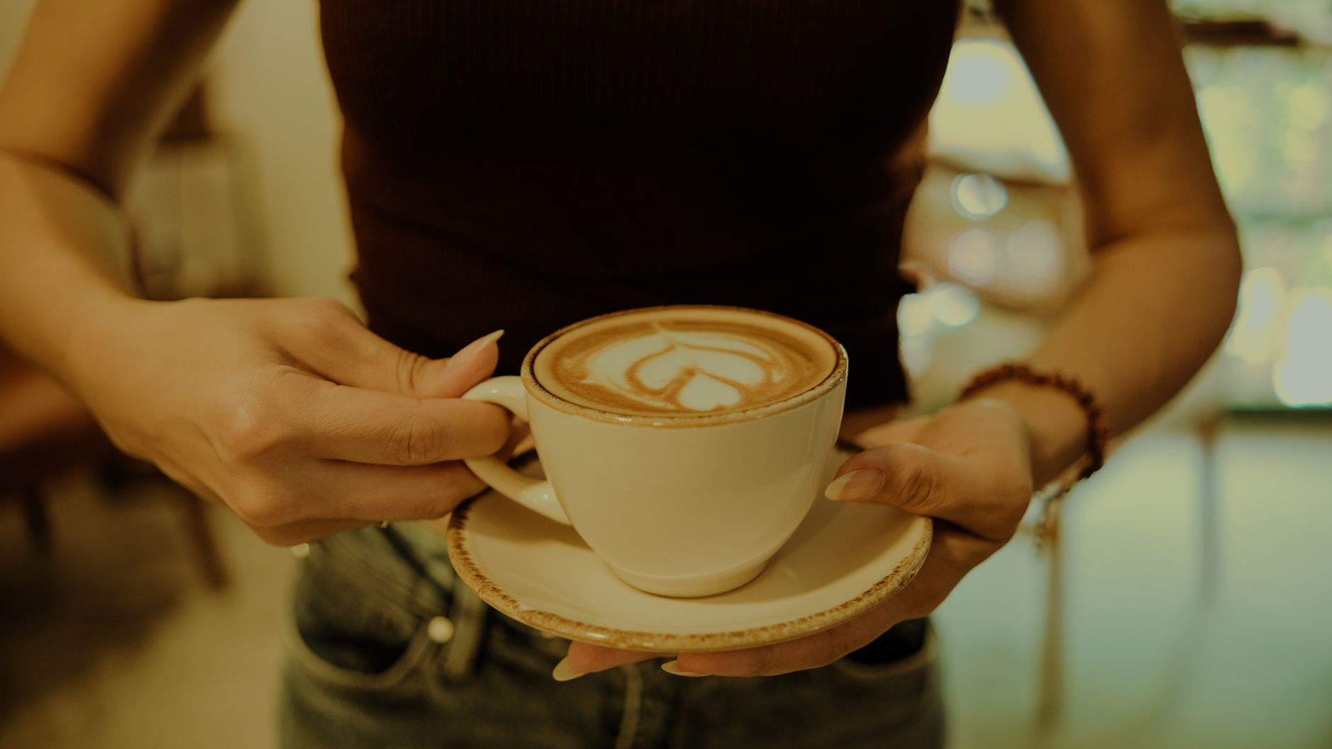 A close-up of hands holding a latte with heart-shaped art in a cozy setting.