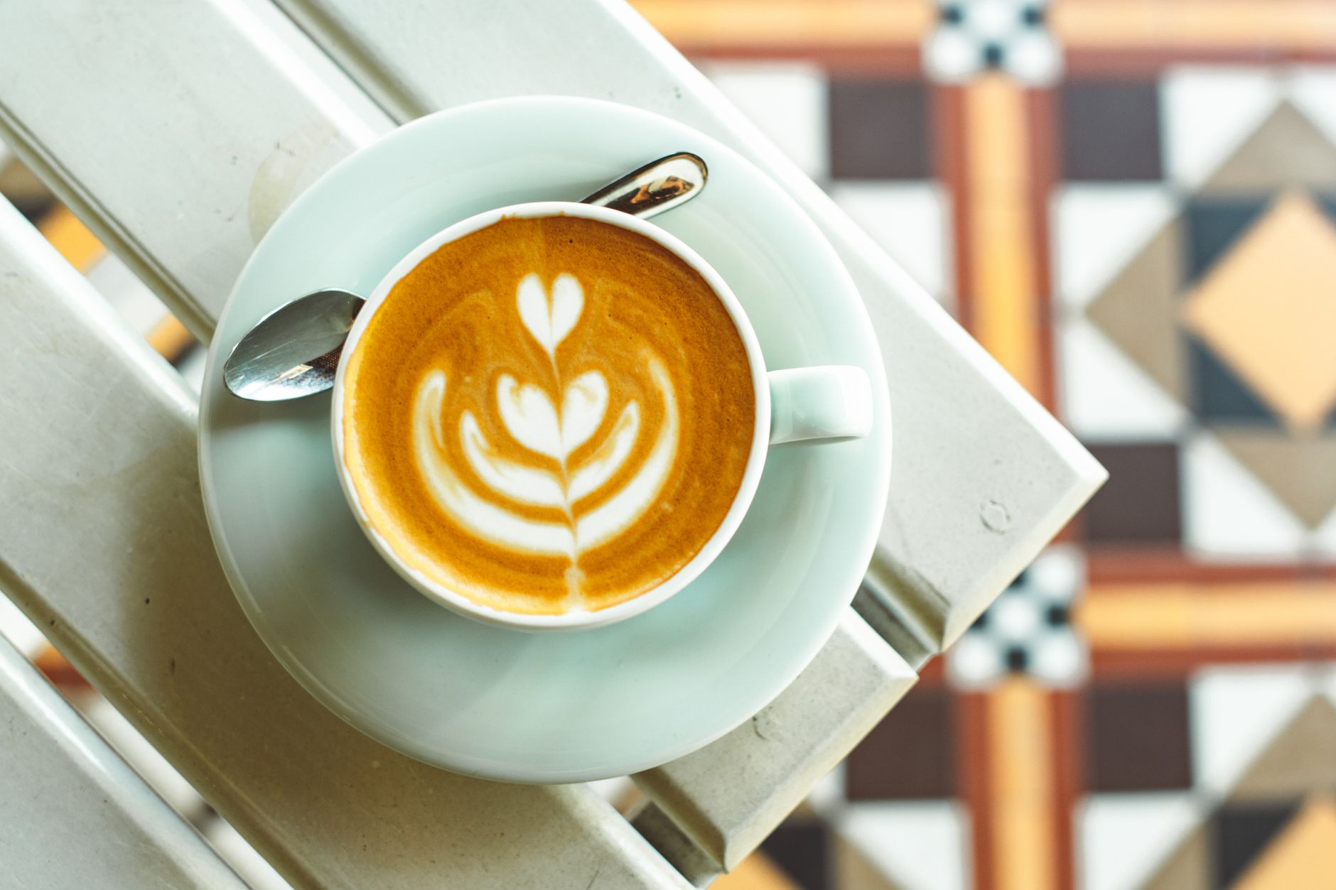 A top-down view of a latte with artistic foam in a white cup placed on a patterned tile surface.