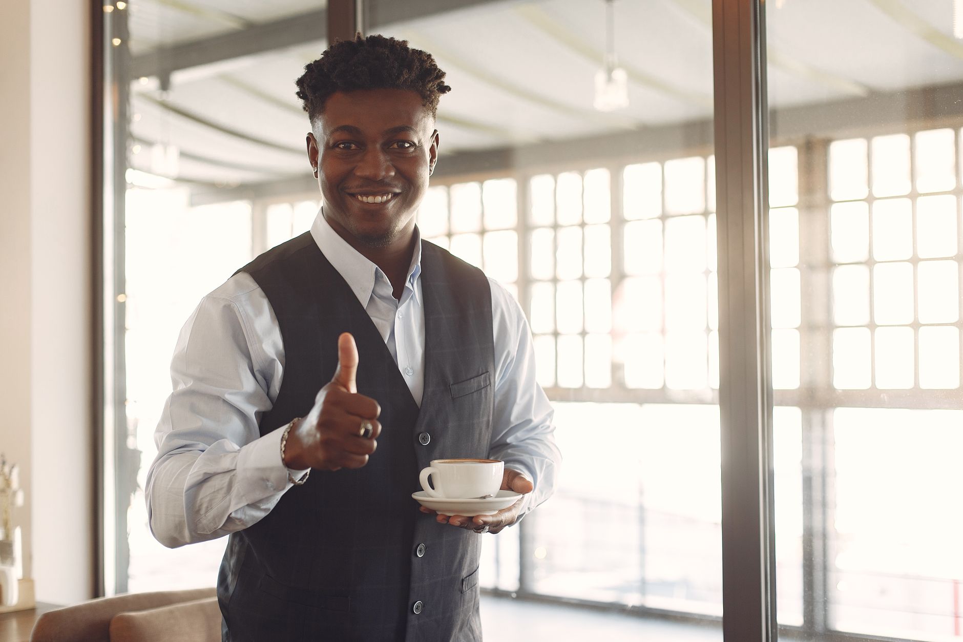 Positive young African American male entrepreneur in formal suit standing with cup of hot coffee in modern cafe near window and showing thumb up while looking at camera