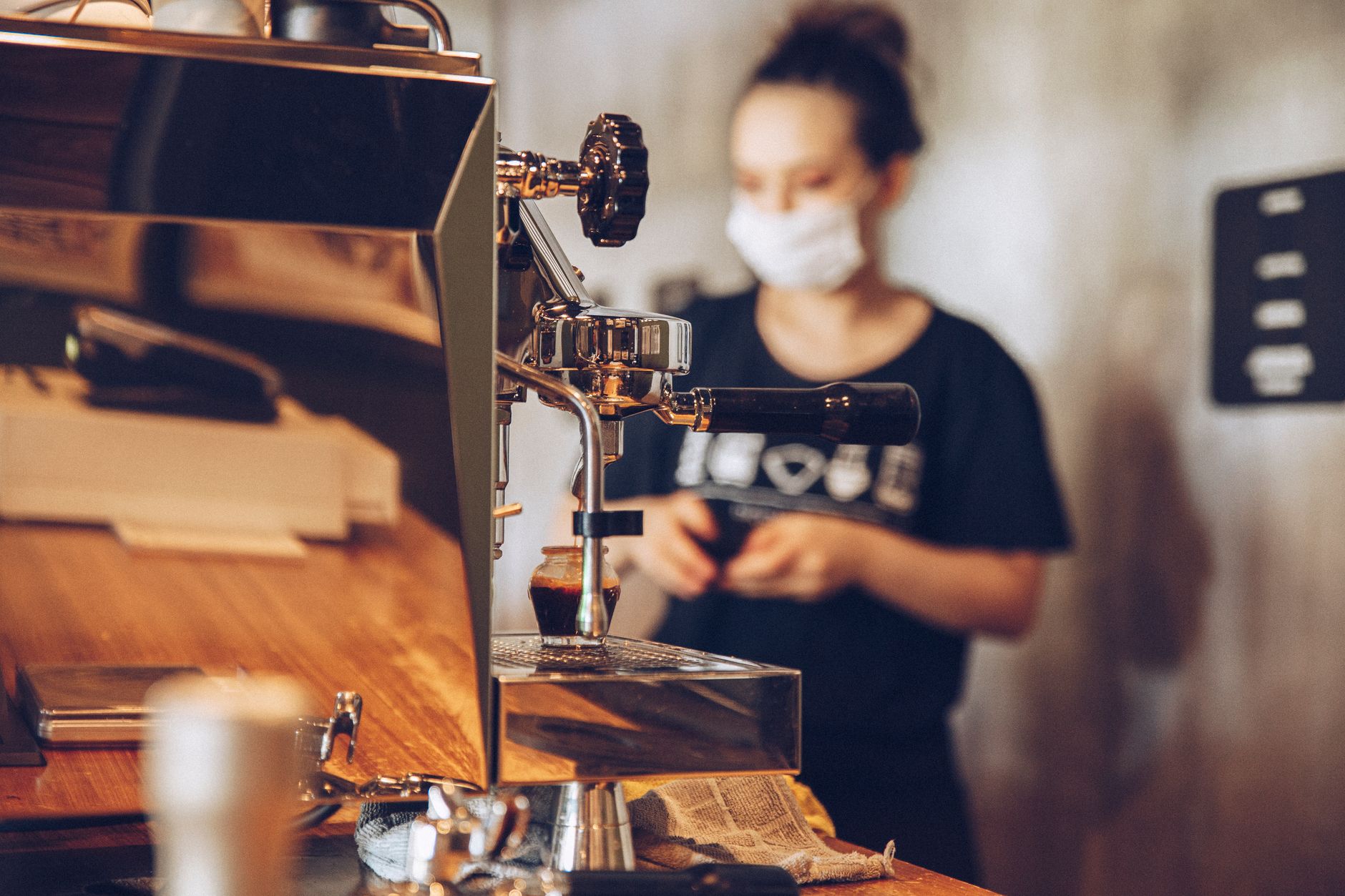 A barista wearing a face mask operates a coffee machine in a trendy cafe setting, focusing on safety and quality coffee-making.