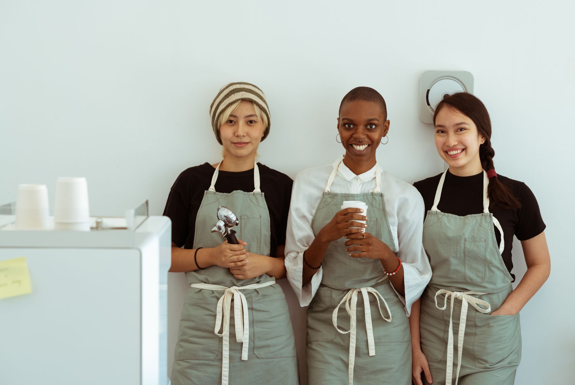 Optimistic smiling diverse female baristas in aprons standing together with portafilter and paper cup against white plain wall and looking at camera happily