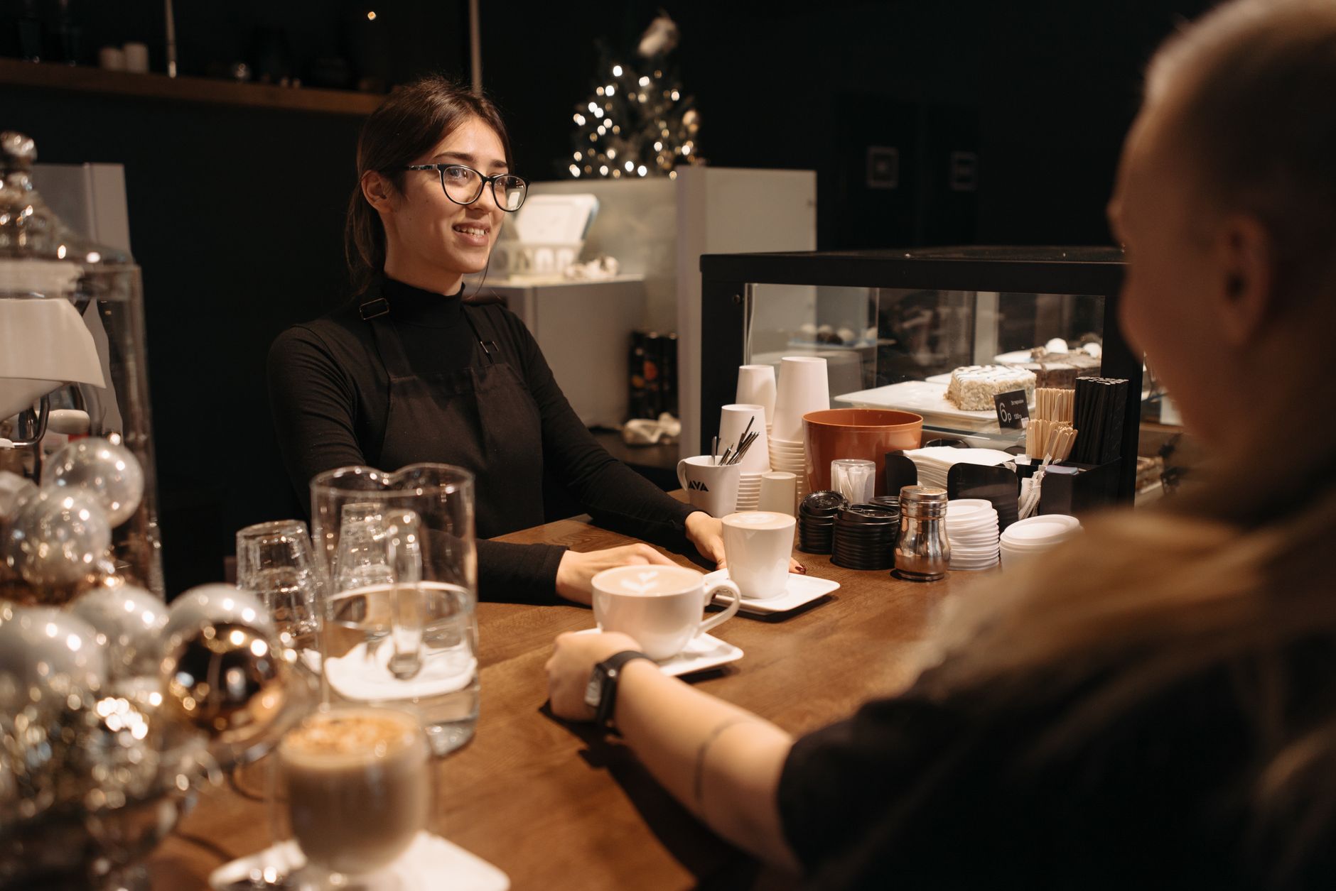 Barista in black apron serves a coffee to a customer at a warm, cozy café setting with festive decor.