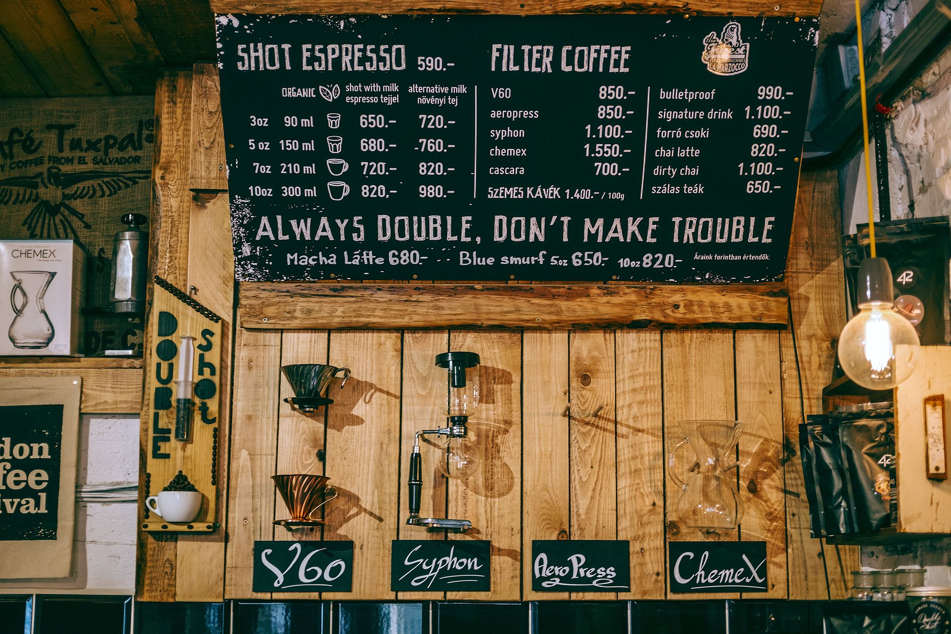 Blackboard with coffee shop menu and Always Double Don t Make Trouble inscription hanging on creative decorated wooden wall with various glassware and brewing tools