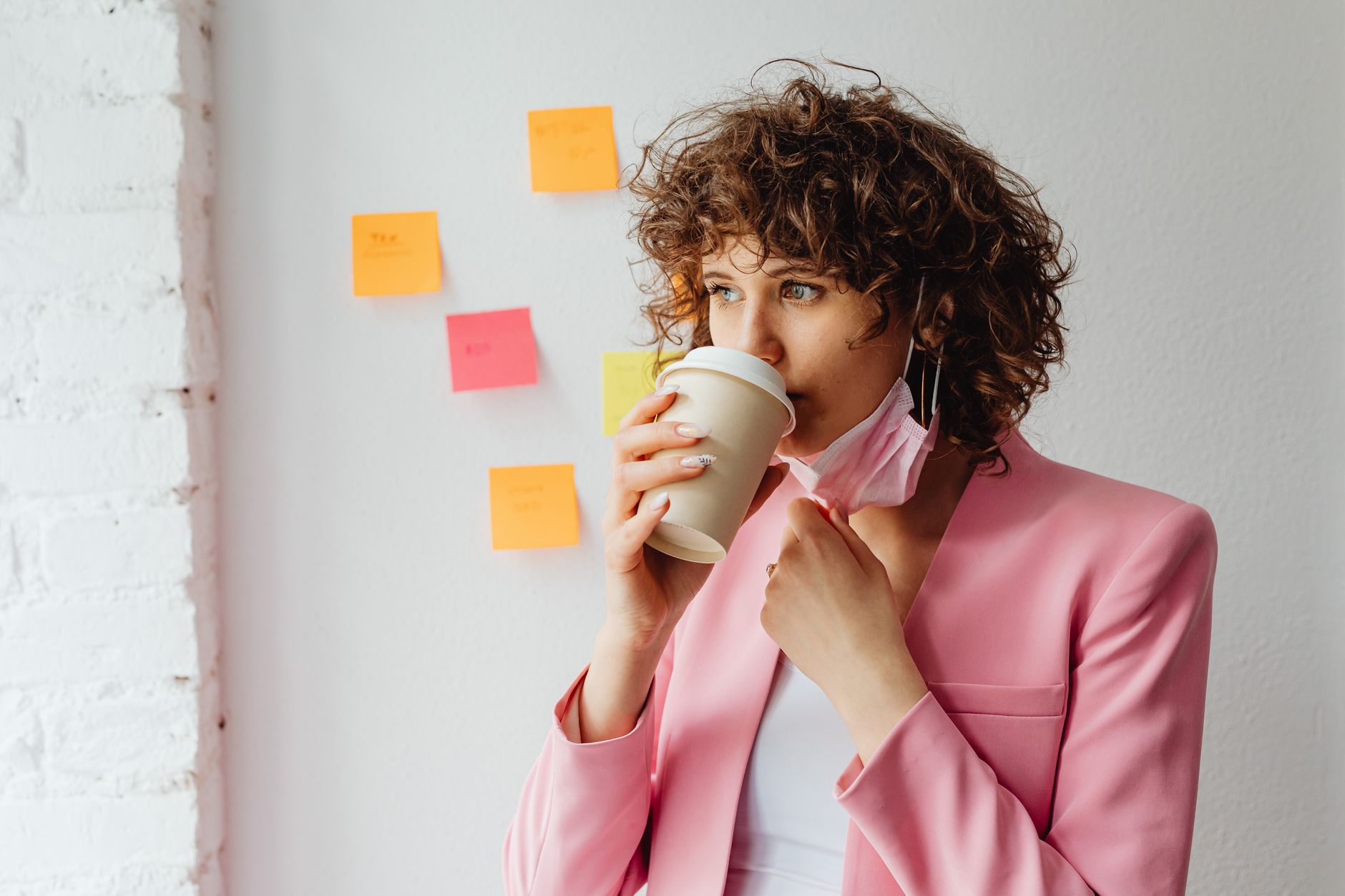 Woman in a pink blazer sipping coffee, holding a face mask, indoors with sticky notes.