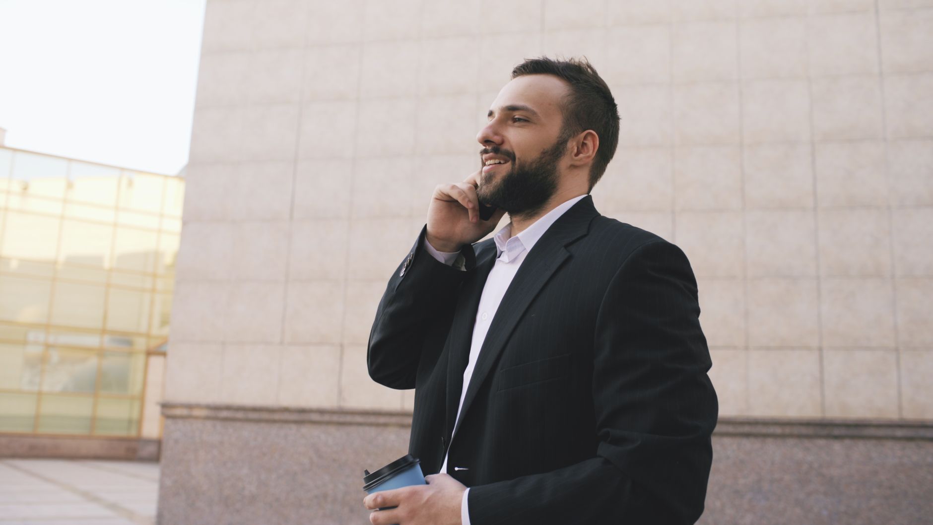 Smiling businessman in suit having a phone call while holding a coffee cup outside an office building.