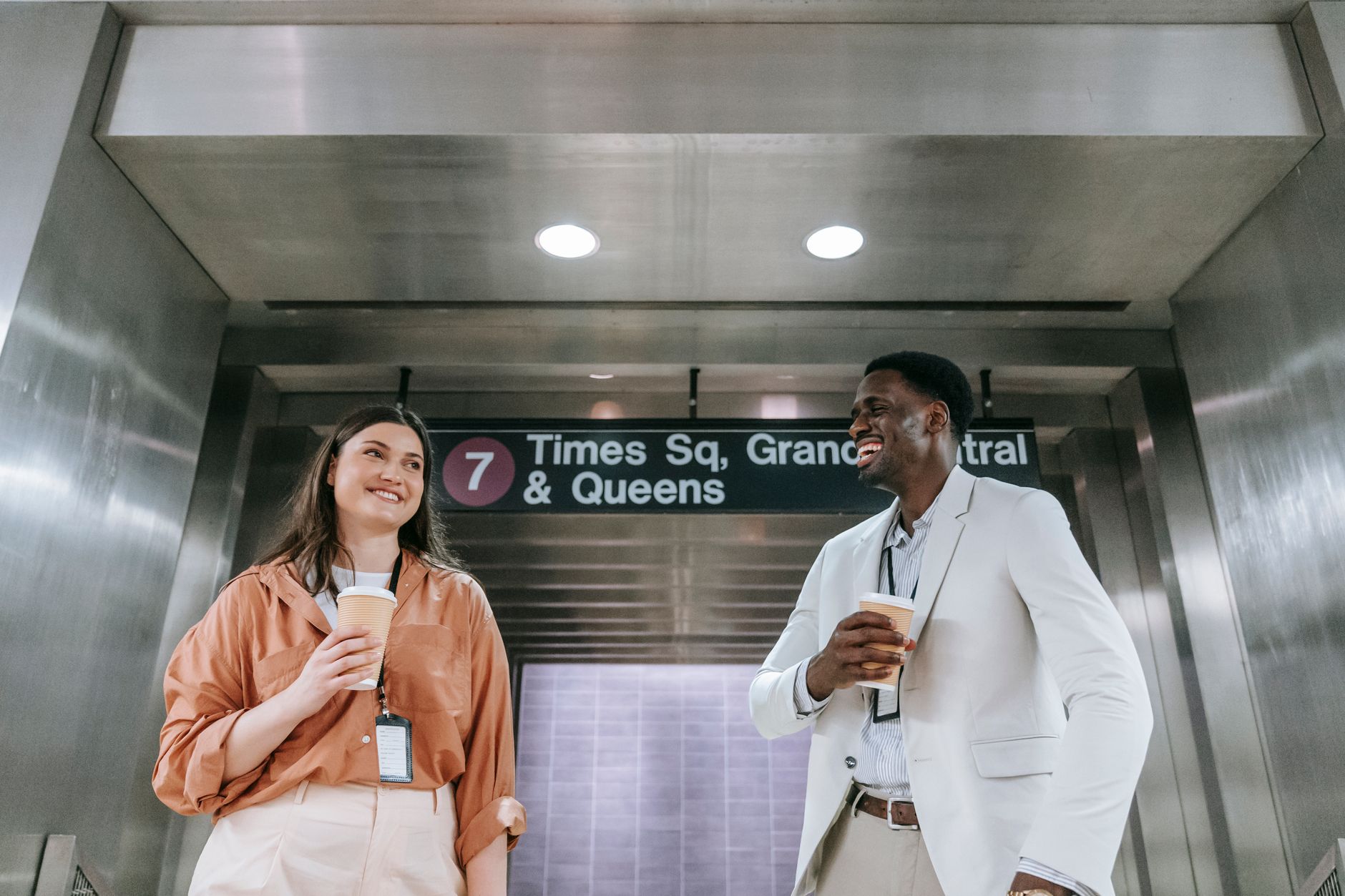 Two colleagues laugh and hold coffee near a subway sign in an underground station.