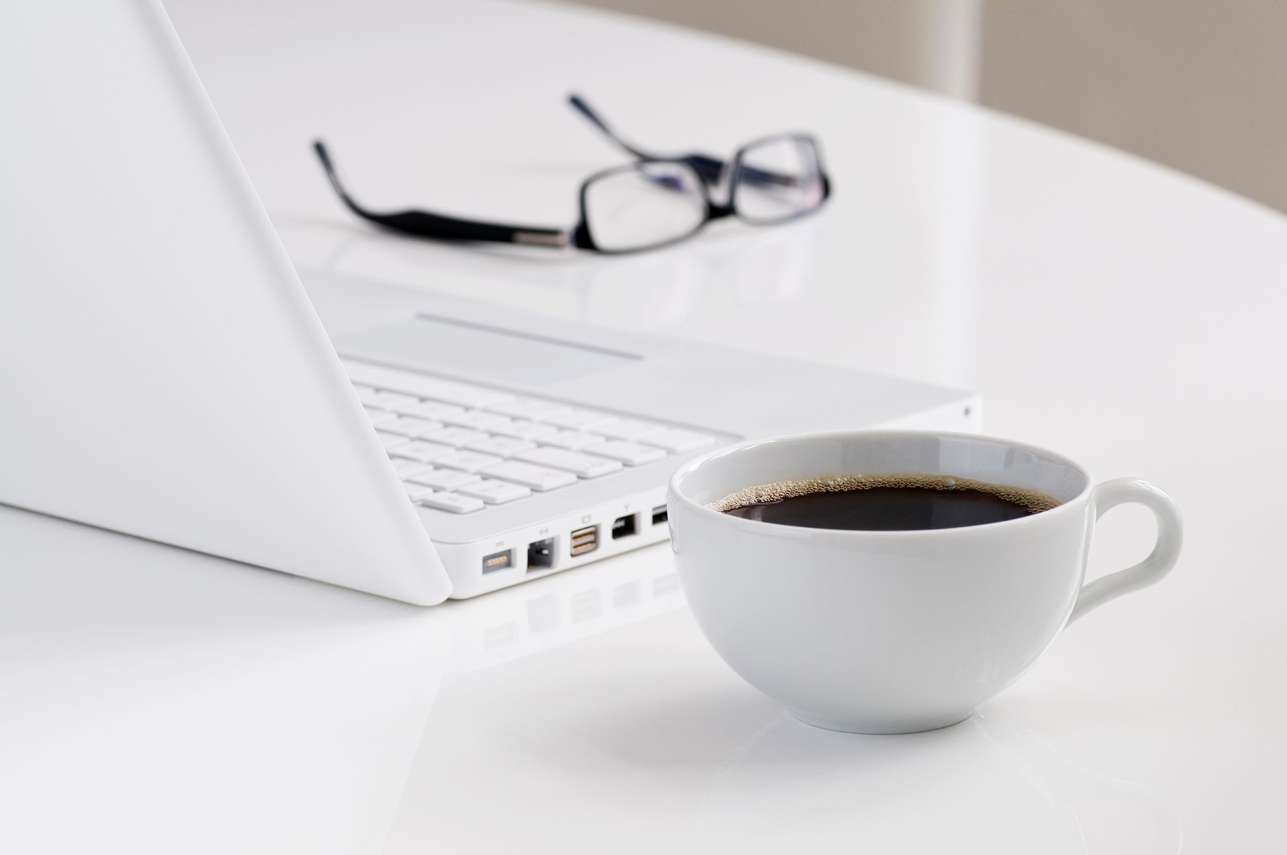 A minimalist workspace featuring a laptop, a cup of black coffee, and eyeglasses on a white desk.
