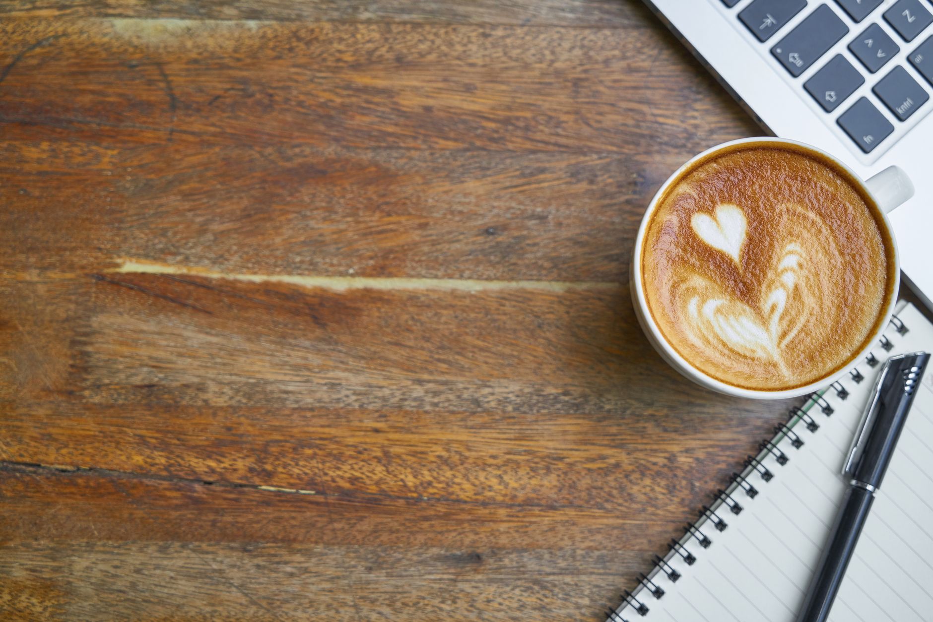 Top view composition with cup of aromatic hot cappuccino placed near modern netbook keyboard and notebook with pen on wooden shabby desk