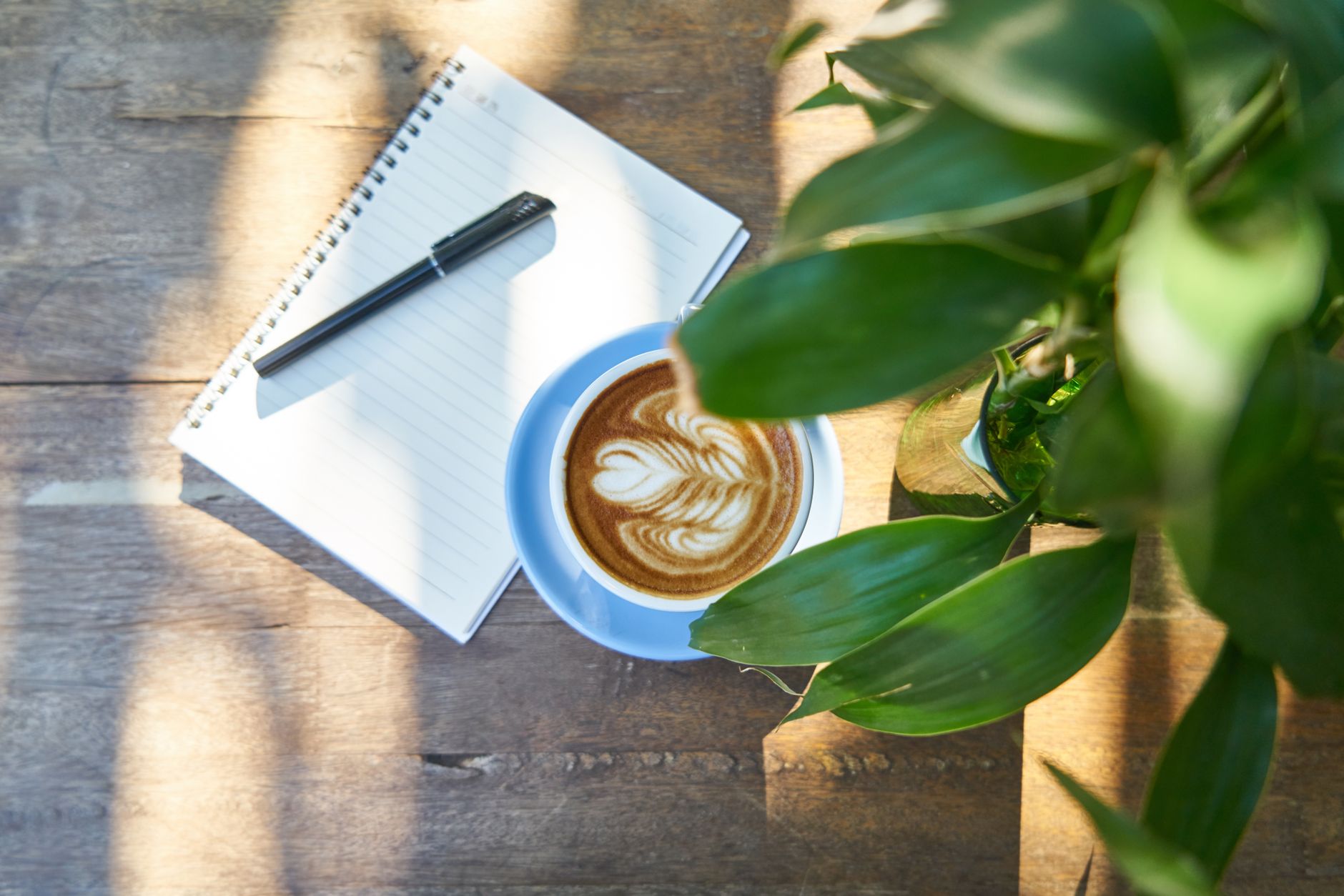 A latte art coffee by a notebook and leafy plant on a wooden table, perfect for inspiration.