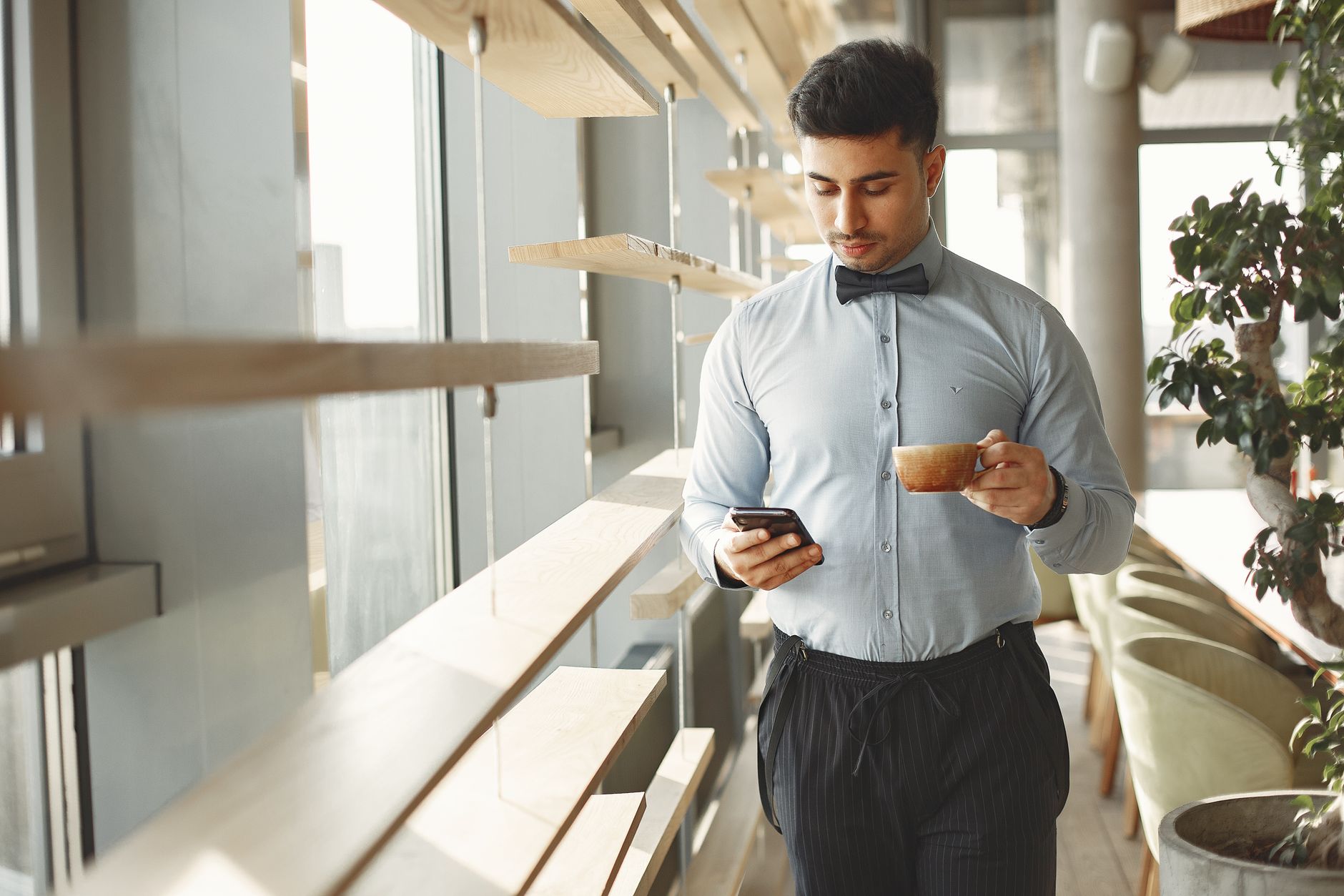Man in business attire holding coffee cup, using smartphone in modern cafe.