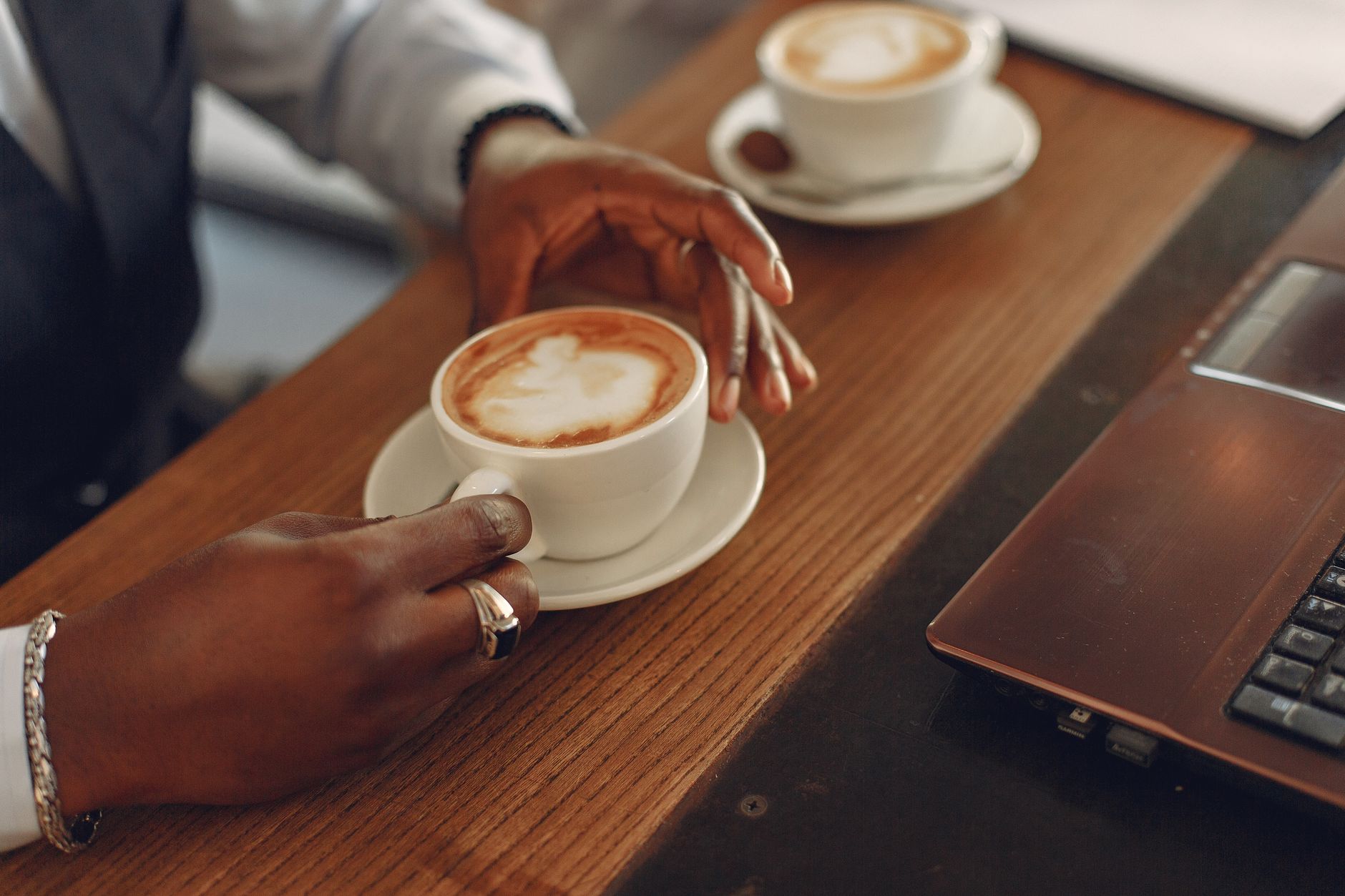 From above view of unrecognizable crop ethnic male entrepreneur in formal clothes drinking fresh coffee while sitting at wooden table near laptop in modern cafe