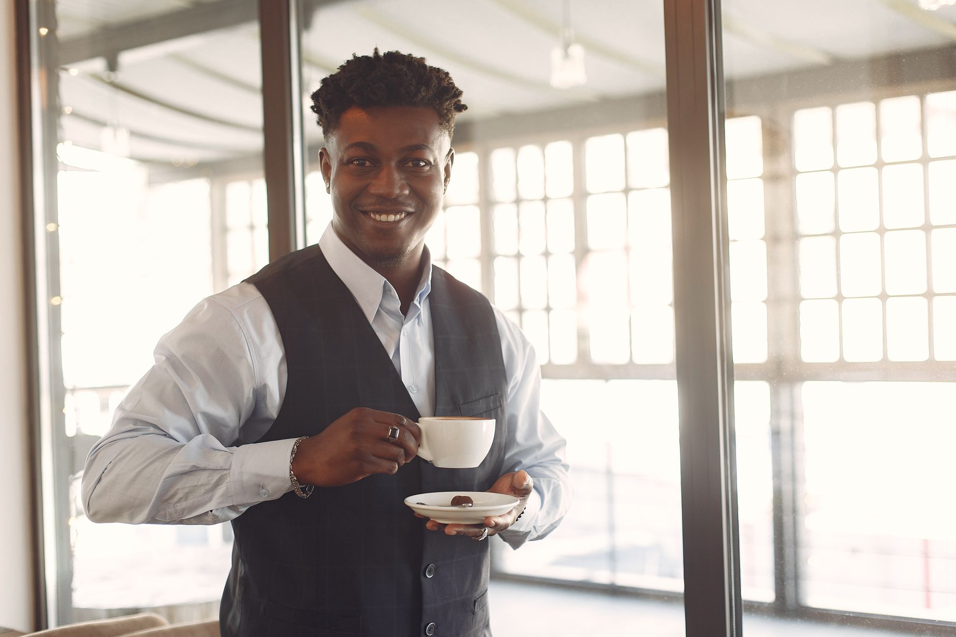 Positive young African American male manager in formal suit standing with cup of fresh coffee near window in light modern cafe and looking at camera