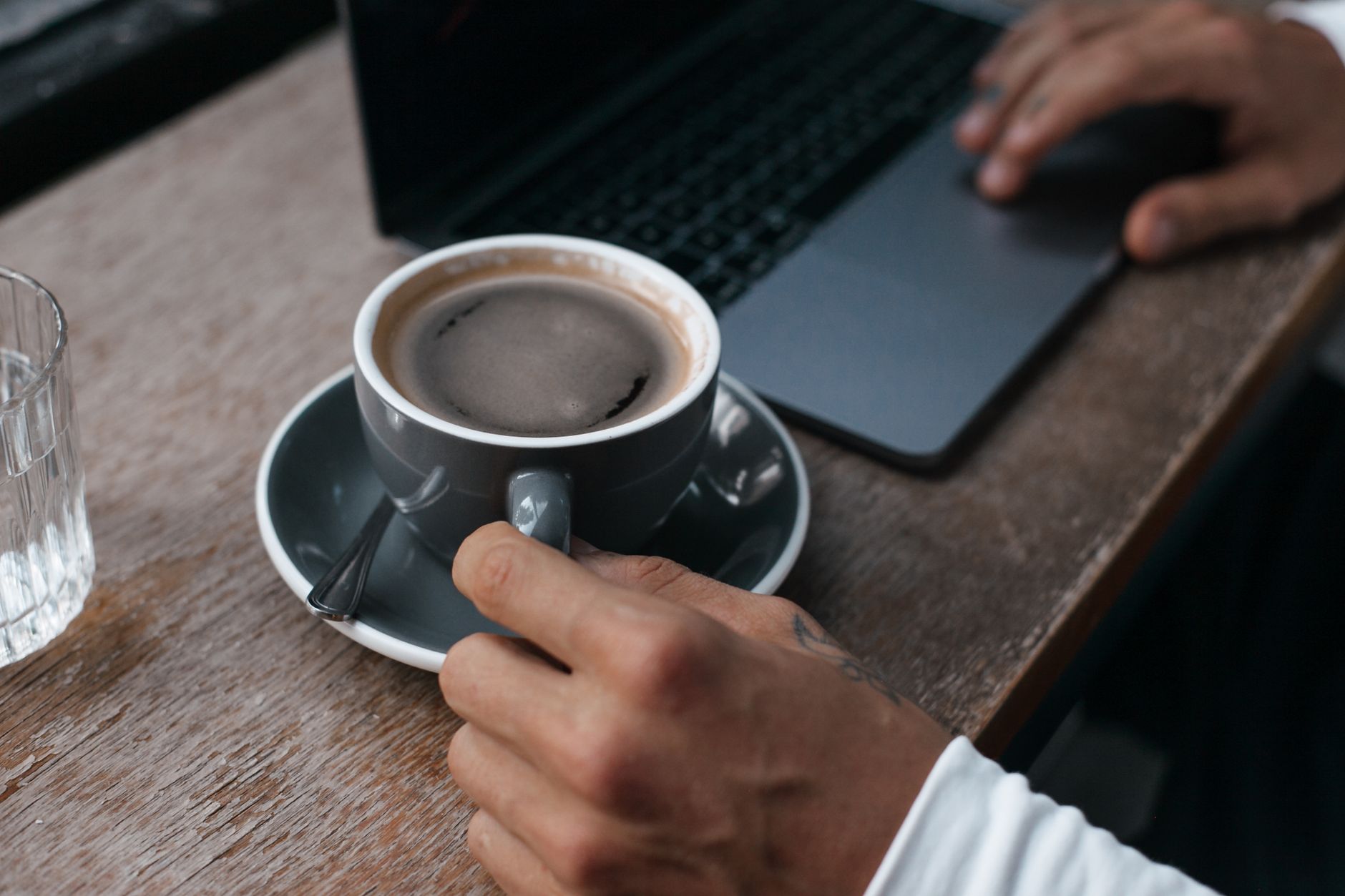 A person holding a coffee cup while working on a laptop at a wooden table.