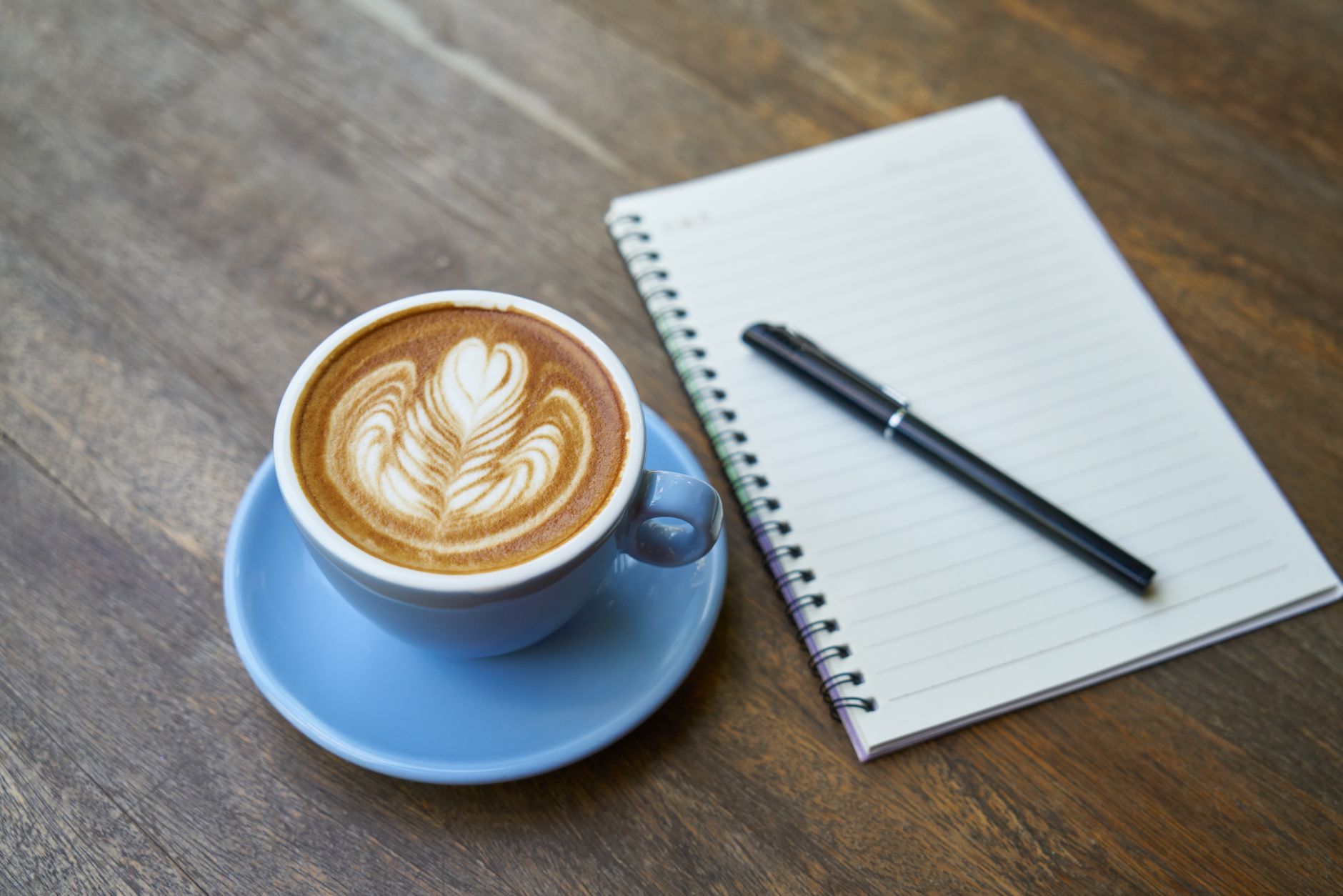 A blue cup of coffee with latte art beside an open notebook and pen on a wooden table.