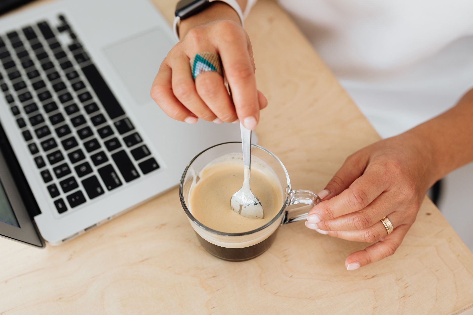 A person stirs coffee in a glass mug beside a laptop on a wooden table.