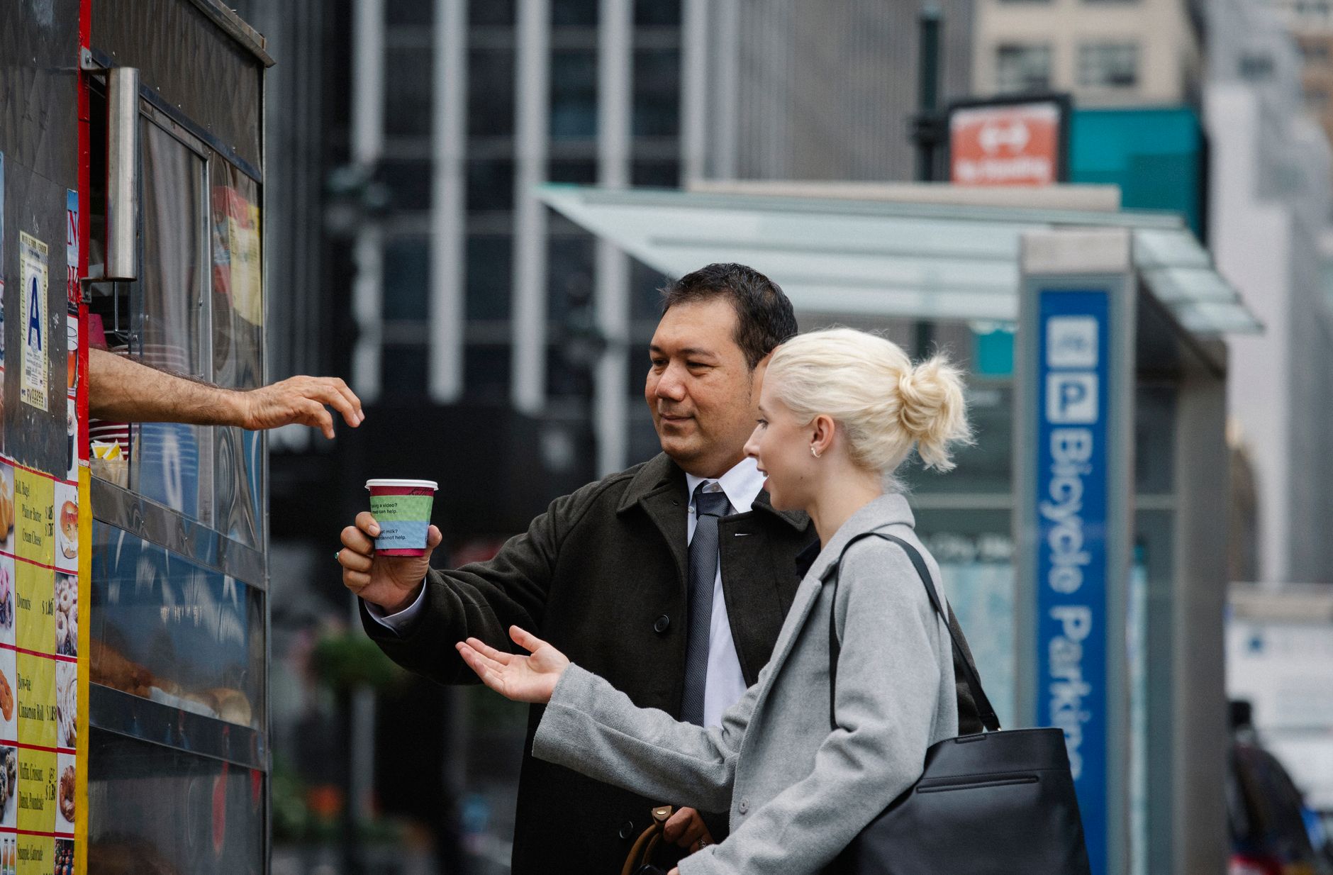 Side view of young female manager with ethnic male colleague in formal clothes buying takeaway coffee from booth on city street during break