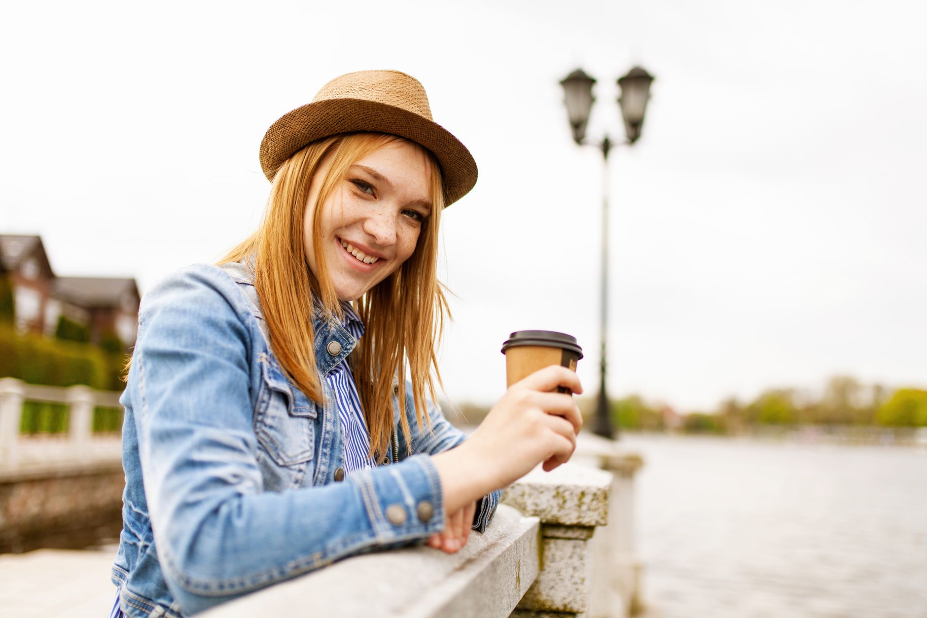 A cheerful woman in a fedora enjoying coffee outdoors by a scenic riverside.