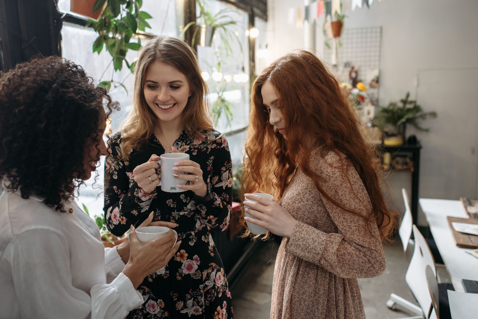 Three women sharing warm beverages in a cozy indoor setting with plants.