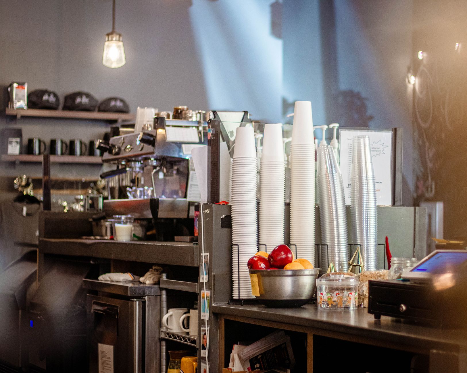 Warm café interior featuring a counter with cups, a bowl of fruit, and an espresso machine.